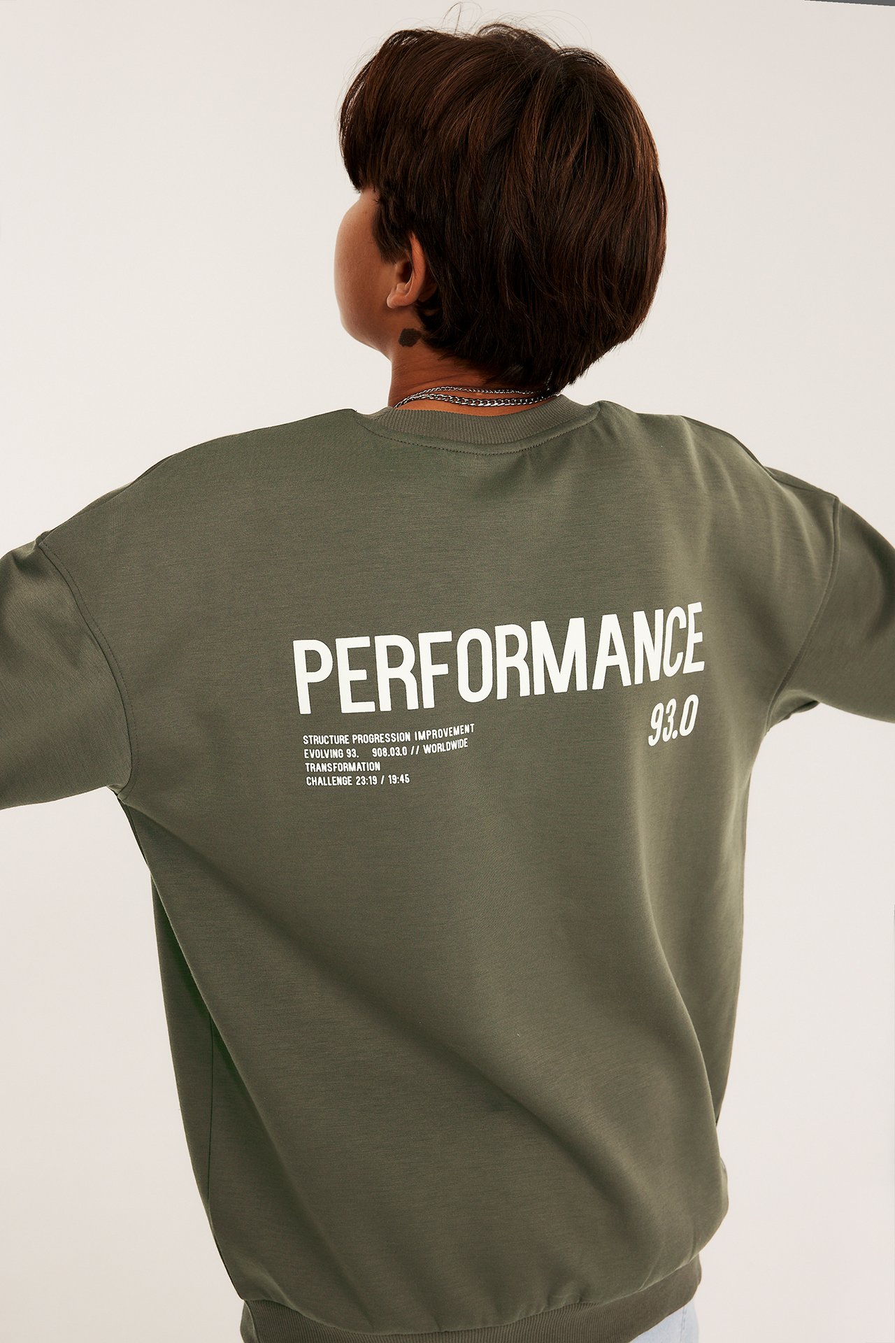 Widok z tyłu: dziecko w luźnej, zielonej bluzie interlock z białym napisem 'PERFORMANCE'.