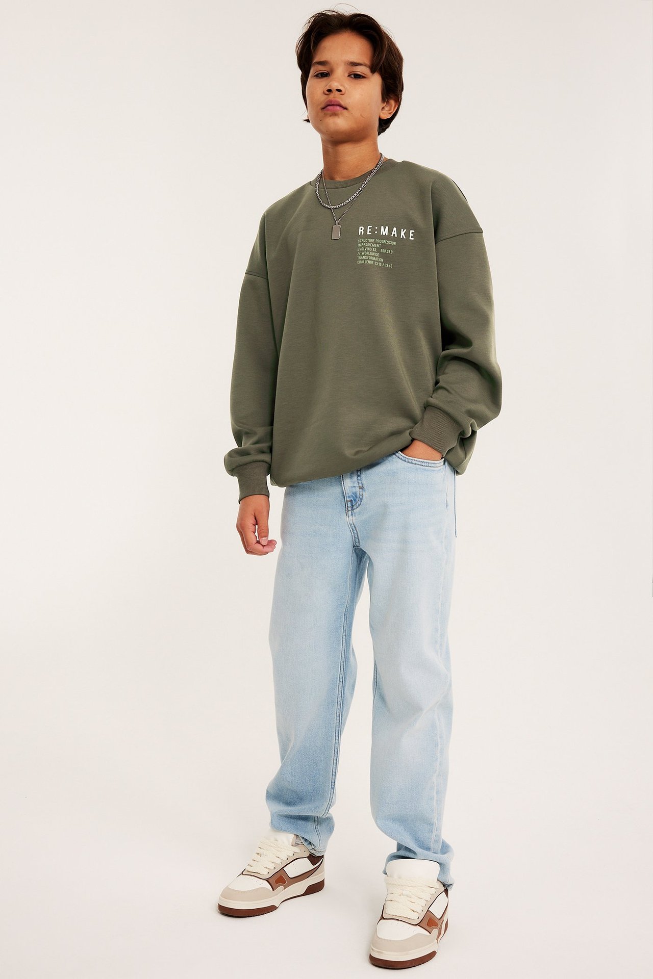 Lapsi vihreässä collegepaidassa rintapainatuksella, hopeakaulakoru, vaaleansiniset farkut ja lenkkarit.