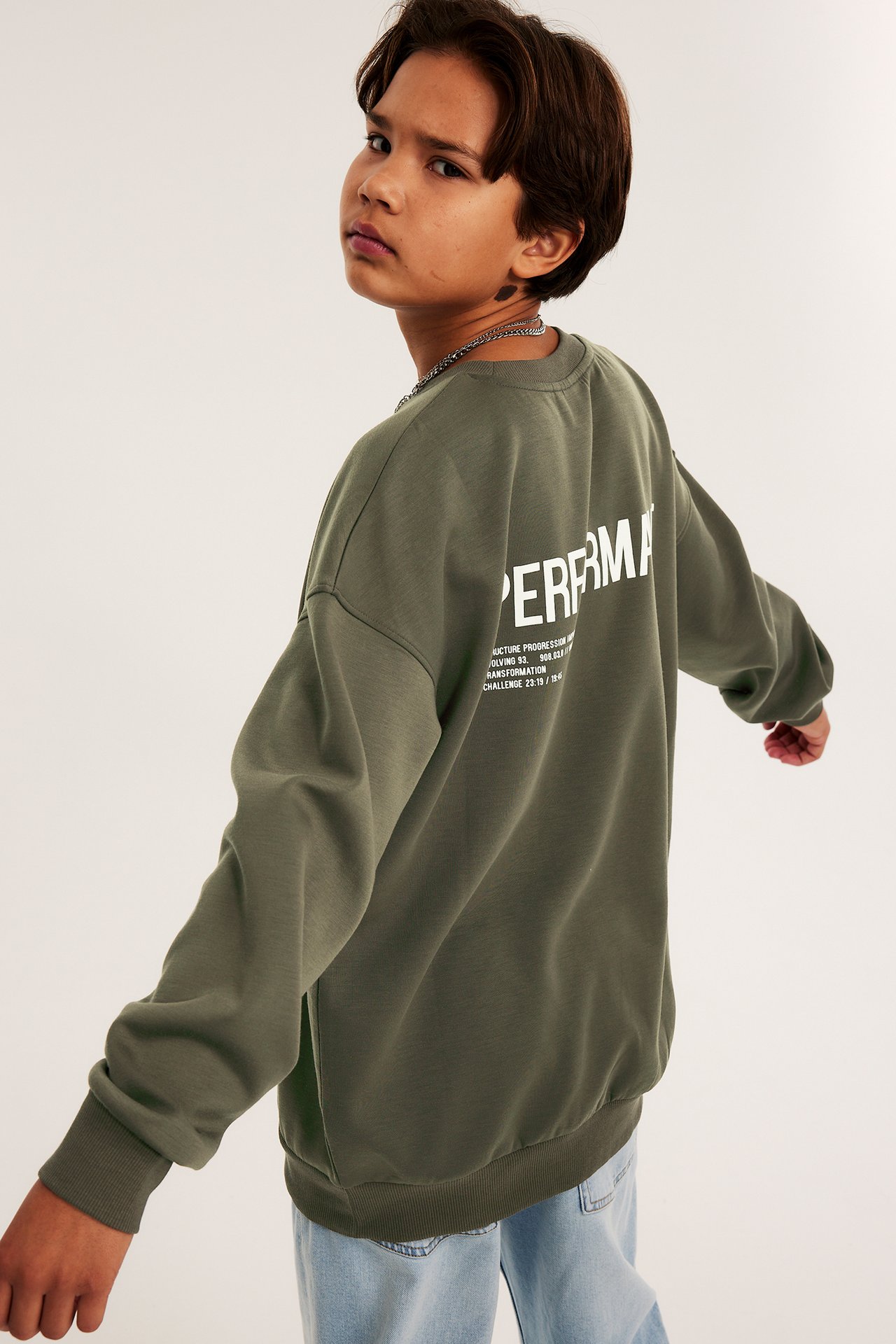 Widok z boku na dziecko w zielonej bluzie interlock z białym nadrukiem na plecach i jasnoniebieskich jeansach.