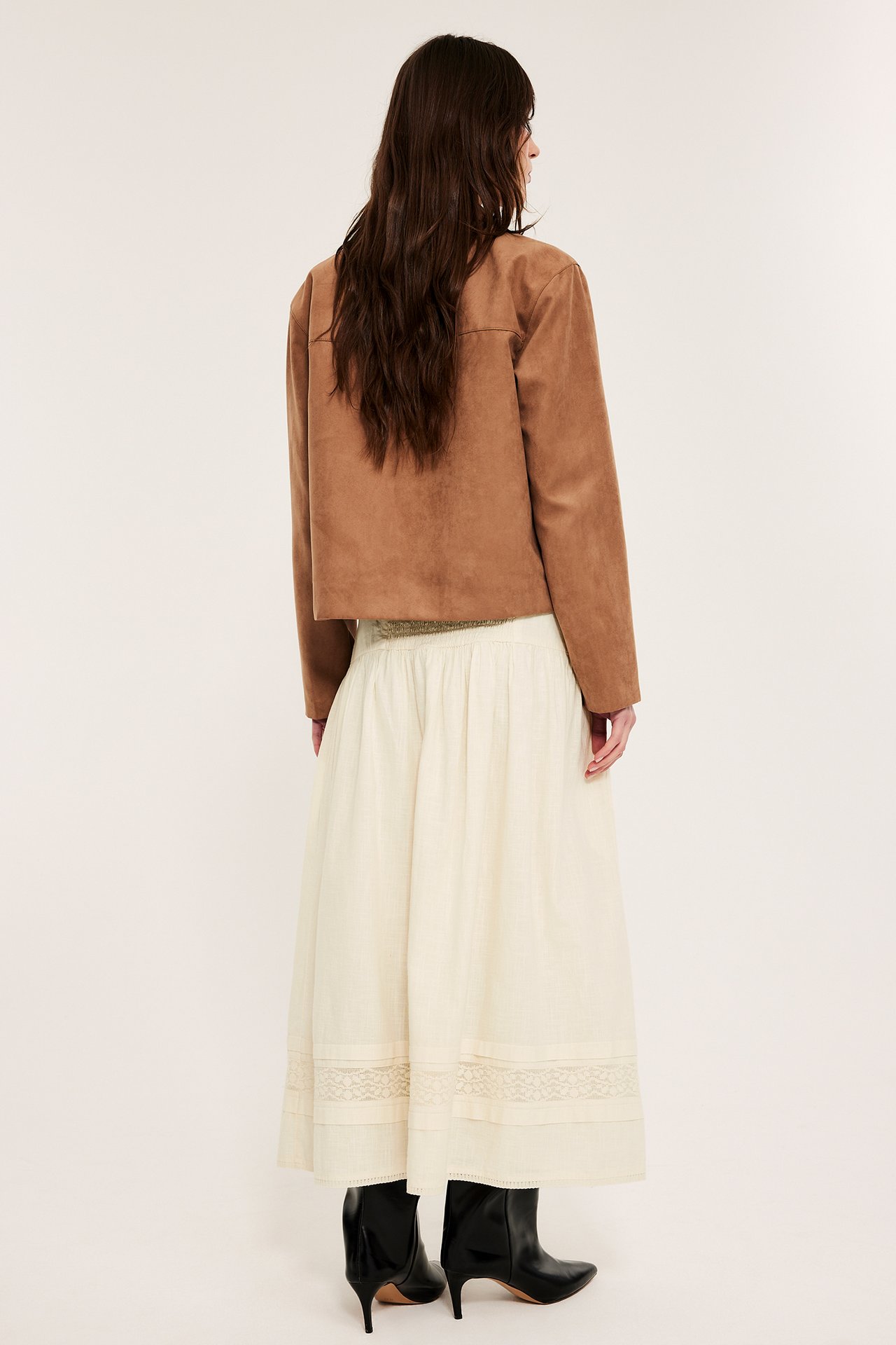 Naisen selkä, yllään luonnonvalkoinen smokkipuuvillahame pitsillä, ruskea lyhyt takki, mustat saappaat.