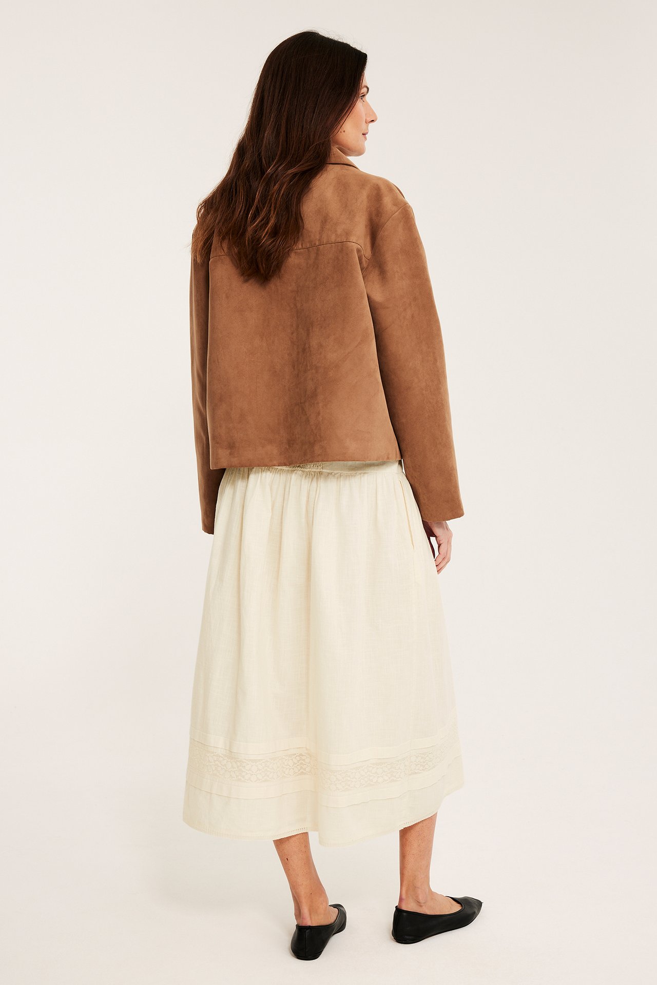 Takakuva naisesta yllään luonnonvalkoinen puuvillahame pitsireunalla, ruskea takki ja mustat ballerinat.