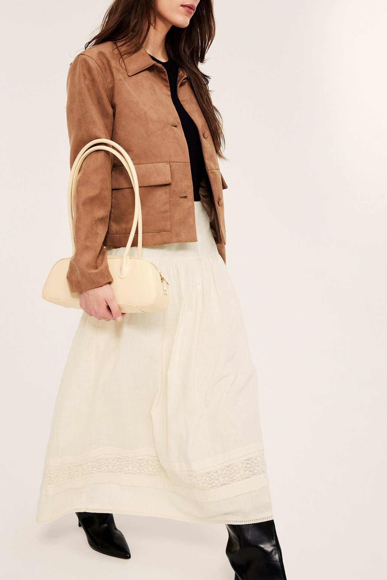 Frontalvisning: Kvinne i offwhite bomullsskjørt med blonder, brun jakke, svart topp, holder kremfarget veske.