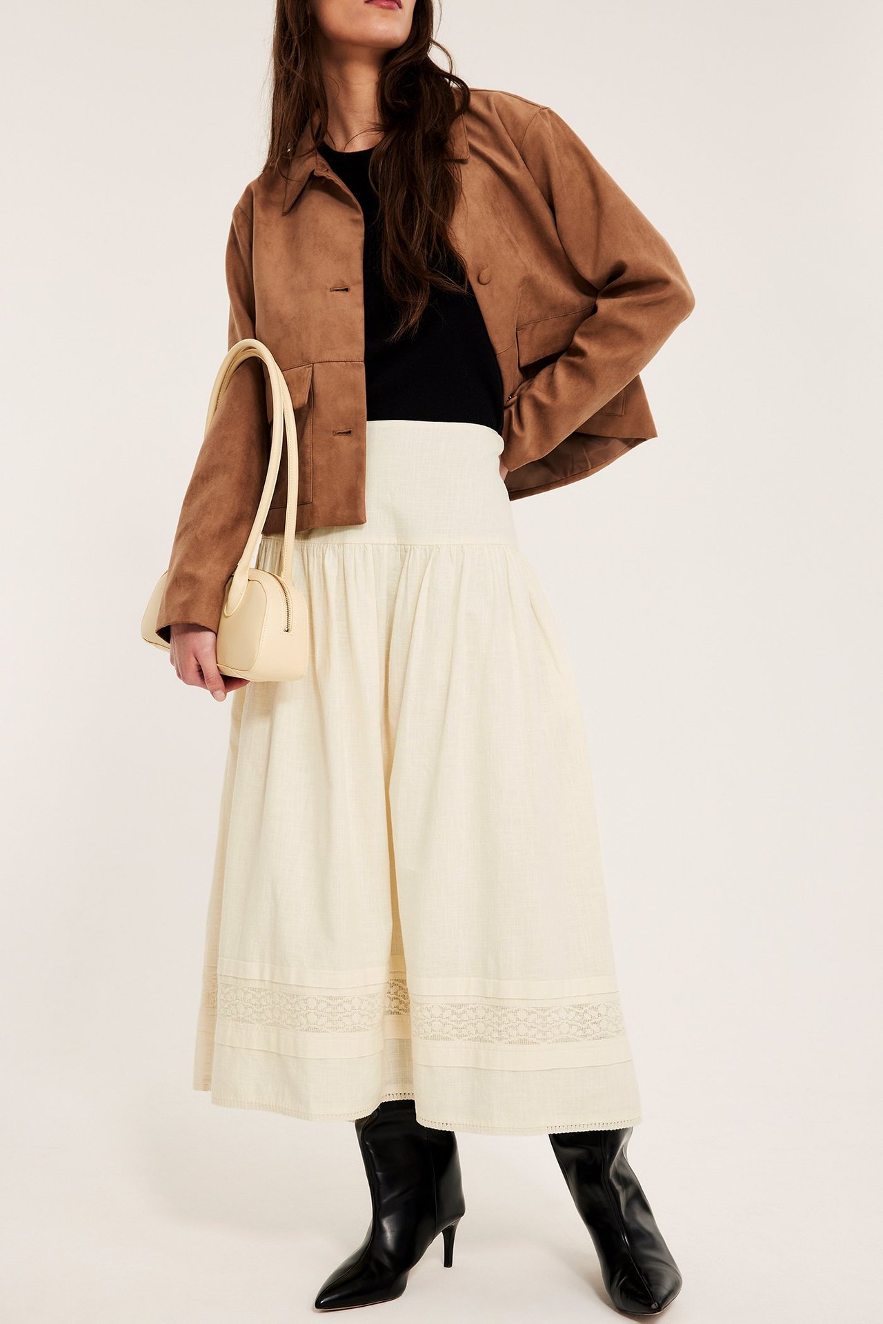 Nainen luonnonvalkoisessa pitsipuuvillahameessa, mustassa topissa, ruskeassa takissa, mustissa saappaissa, pitäen beigeä laukkua.