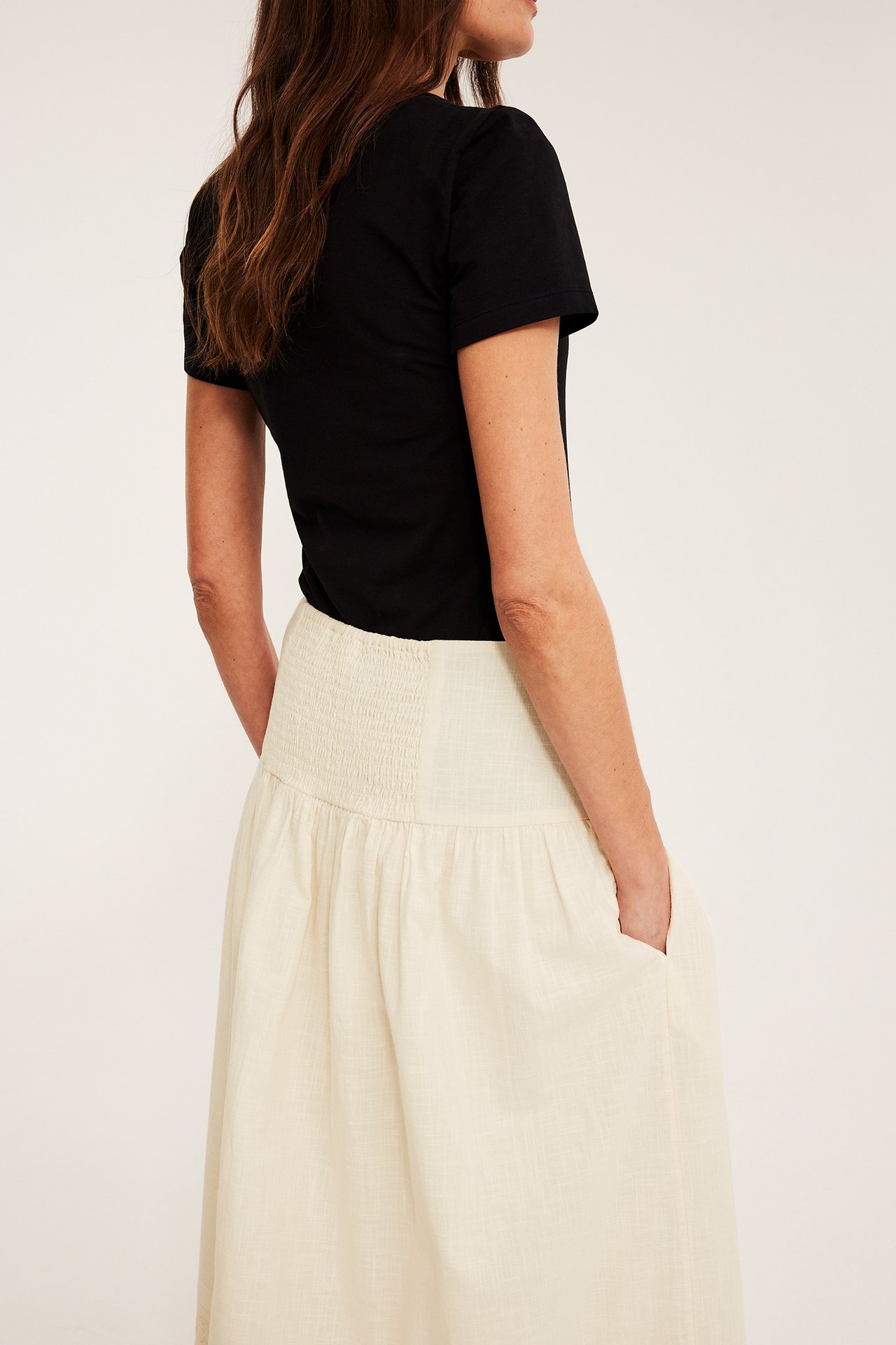 Sivulta-takaapäin: nainen luonnonvalkoisessa puuvillahameessa, smokkirypytyksellä, taskuilla ja mustassa t-paidassa.