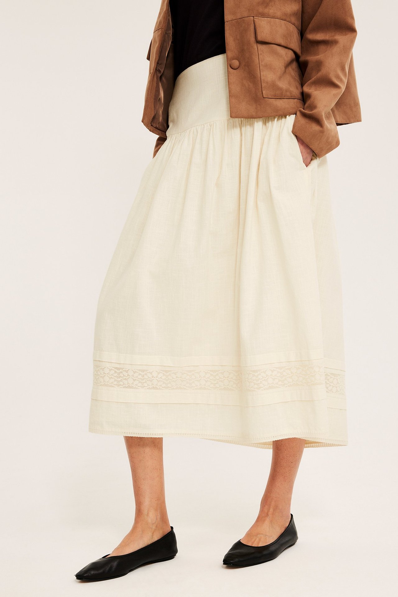 Frontvisning: kvinne i offwhite bomullsmidi-skjørt med blonder, brun jakke og svarte ballerinasko.