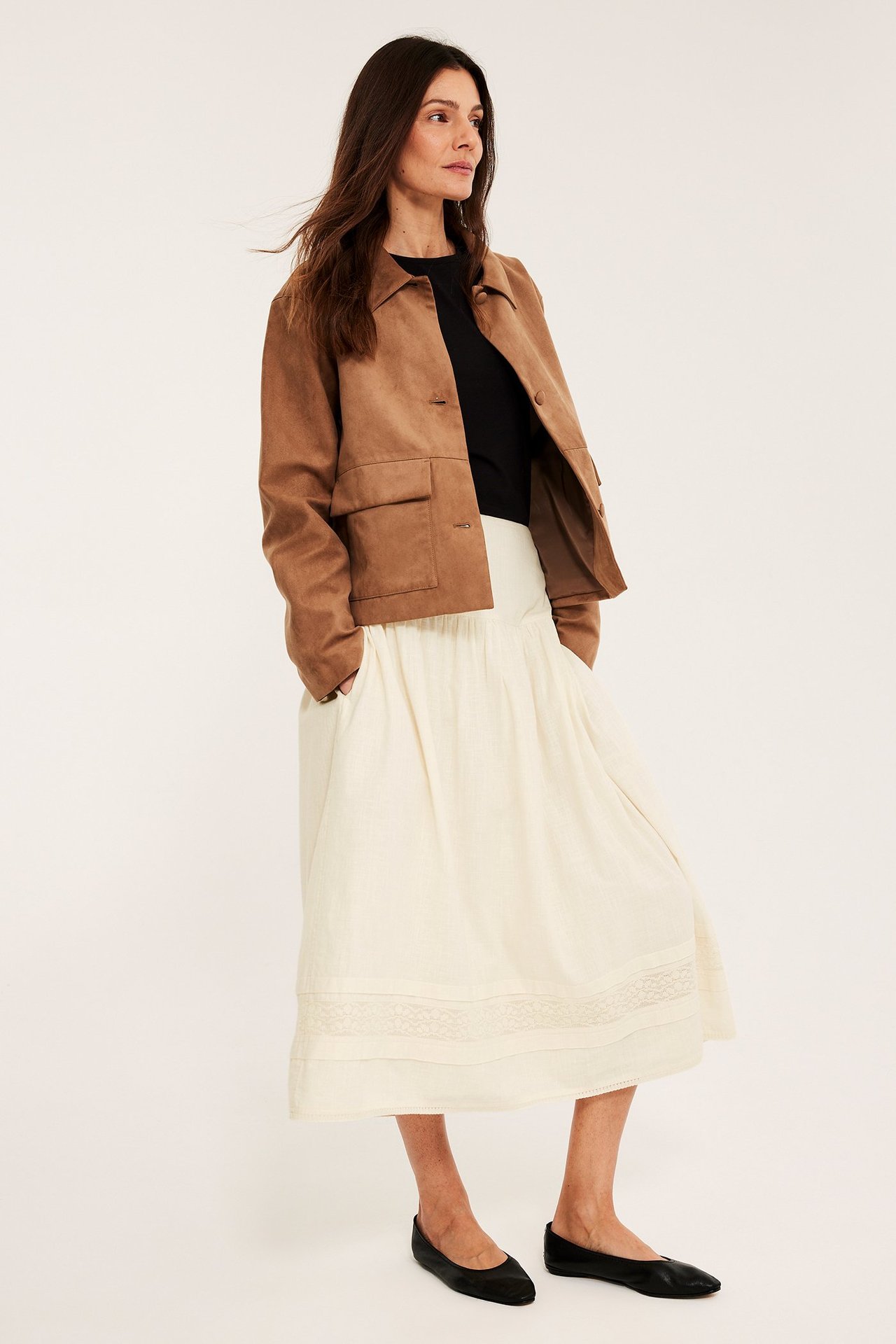 Kvinne i offwhite bomullsskjørt med blonder, brun jakke, svart t-skjorte og svarte ballerinasko.