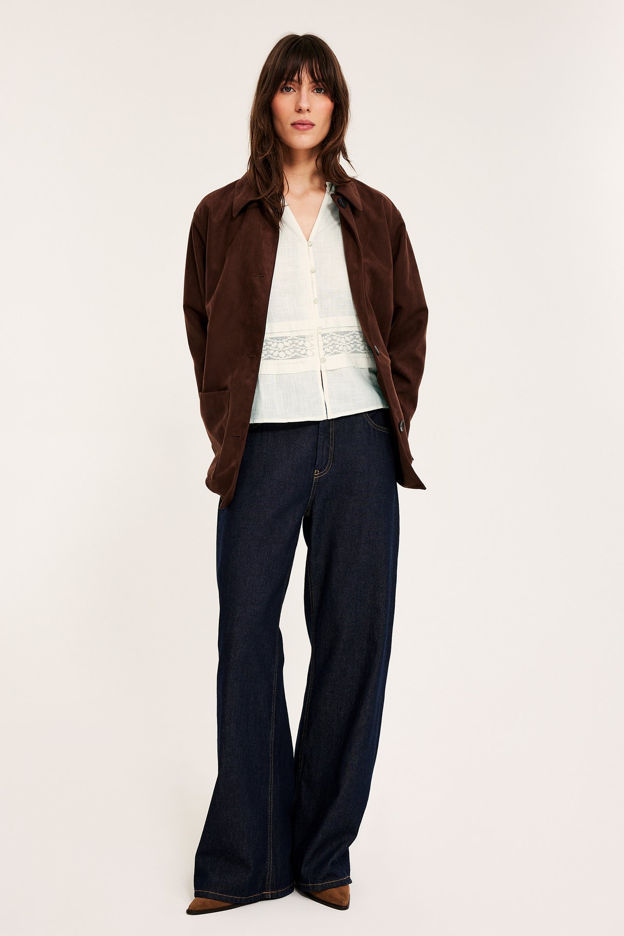 Frontutsikt: kvinne i offwhite bomullsbluse med blonder, brun jakke og mørkeblå vide jeans.