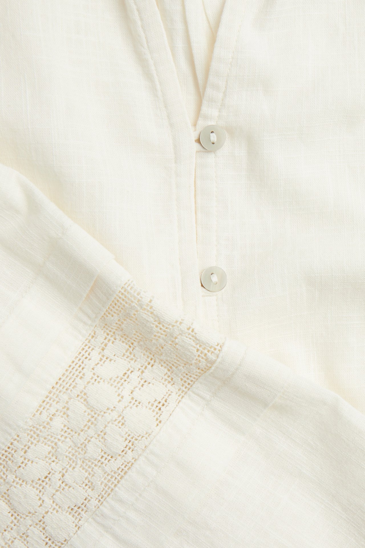 Närbild av en offwhite bomullsblus med V-ringning, ljusa knappar och mönstrad spets på ärmen.