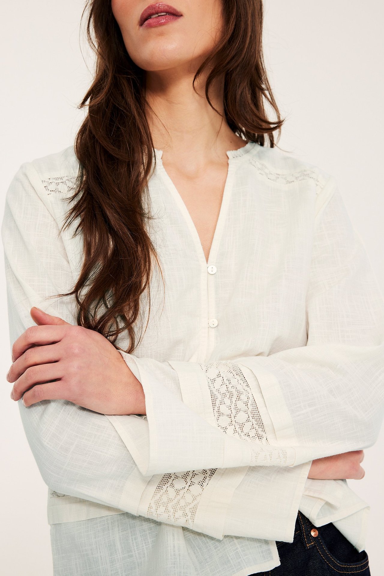 Zbliżenie, przód: Kobieta w kremowej bawełnianej bluzce z koronką, guzikami, na ciemnych dżinsach.