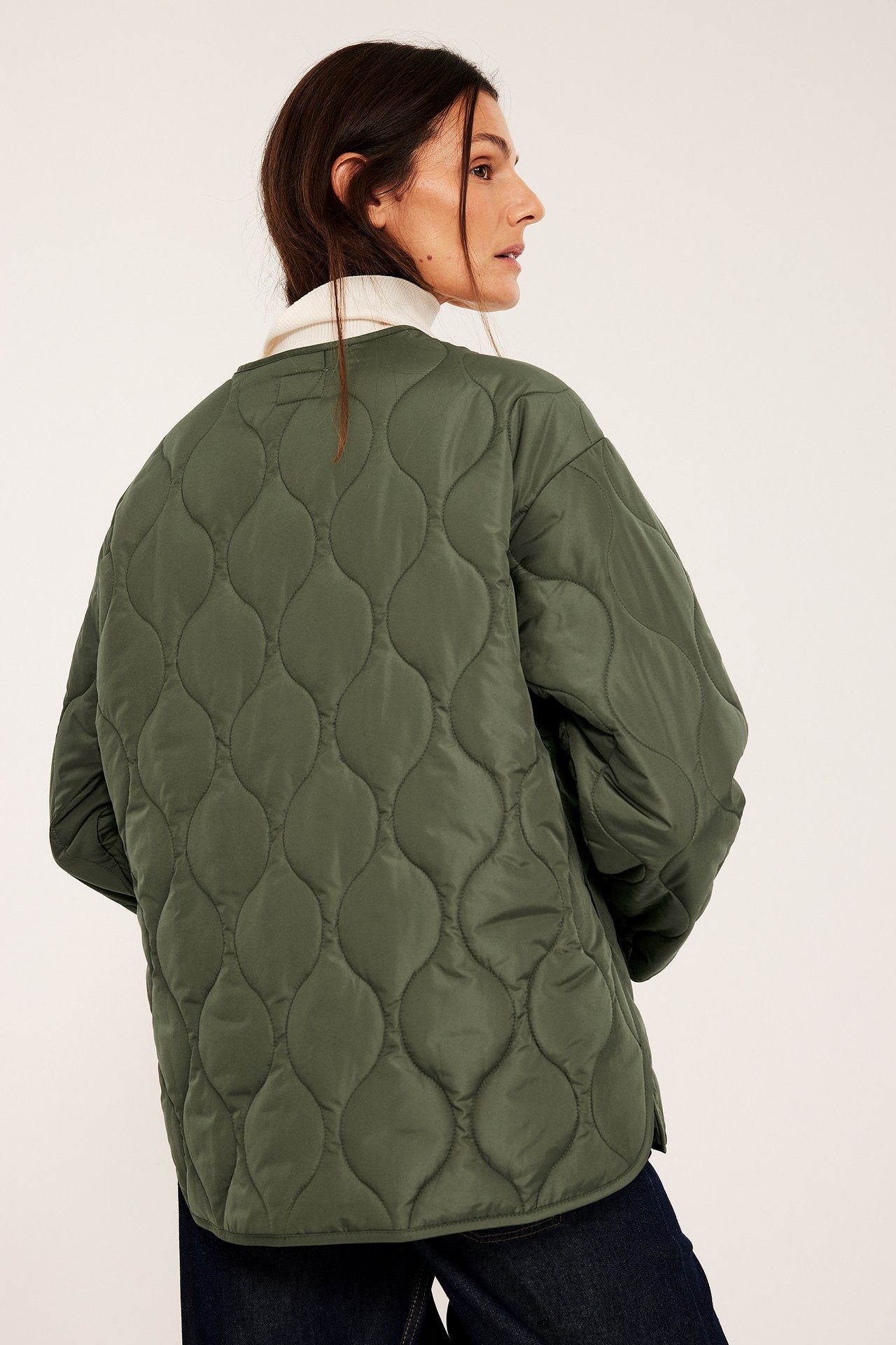 Bakifrån: Kvinna i oversized olivgrön quiltad jacka över krämvit polotröja och mörkblå jeans.