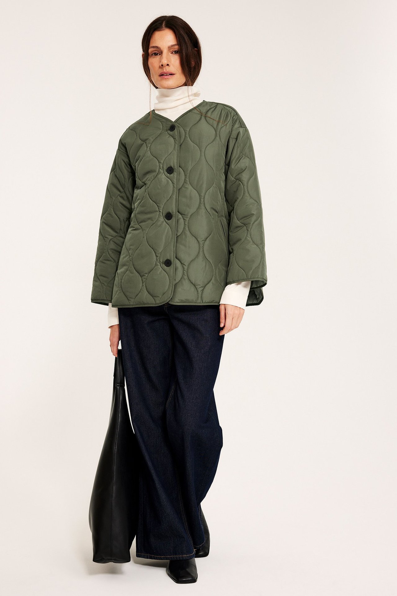 Framsida: Kvinna bär grön quiltad jacka med svarta knappar, vit polotröja, mörkblå jeans, håller svart väska.