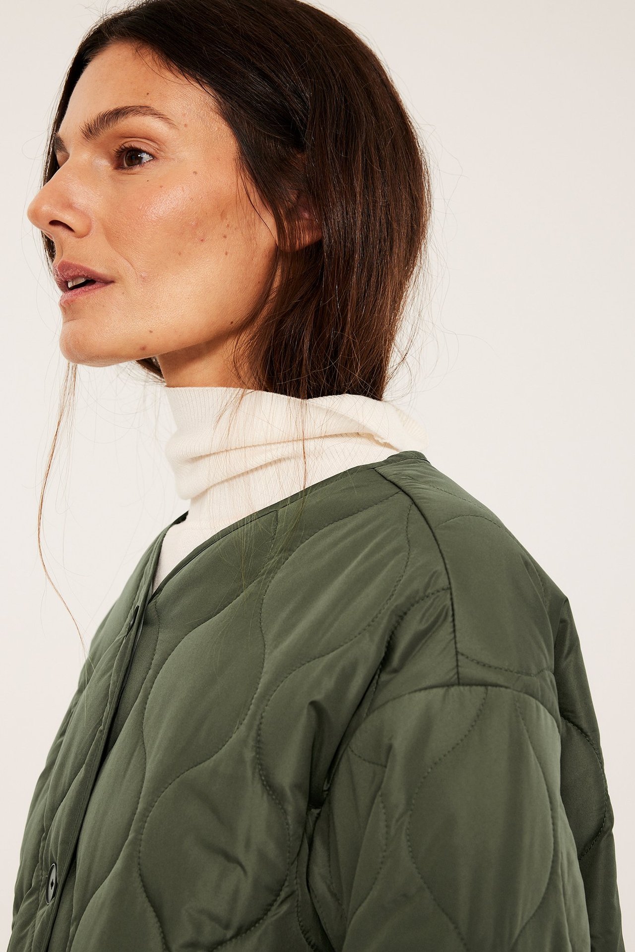 Nærbilde, sideblikk: Kvinne i grønn vattert jakke med mørke knapper over en kremfarget høyhalset genser.