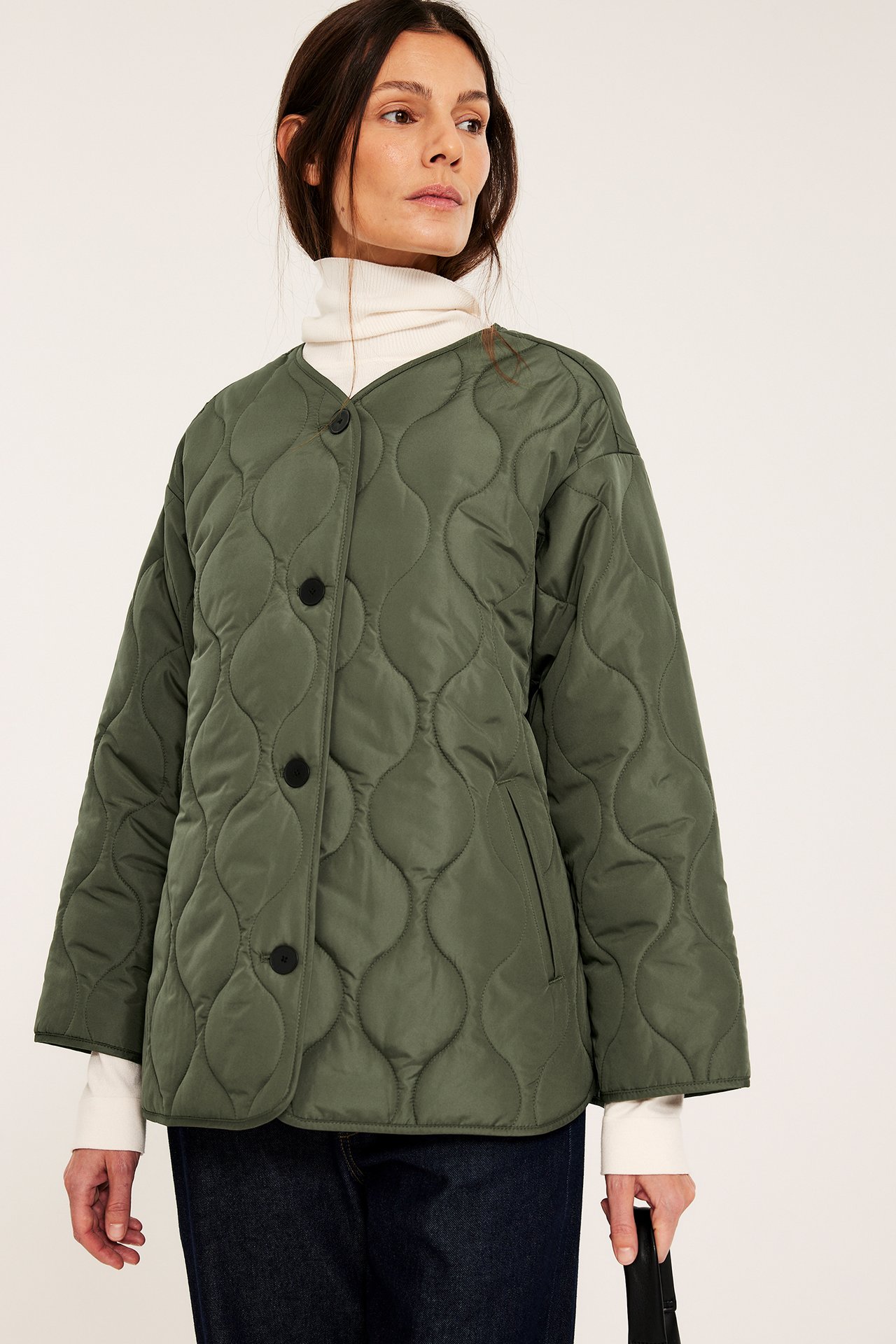 Kvinna bär en grön quiltad jacka med svarta knappar över en vit polotröja och jeans.