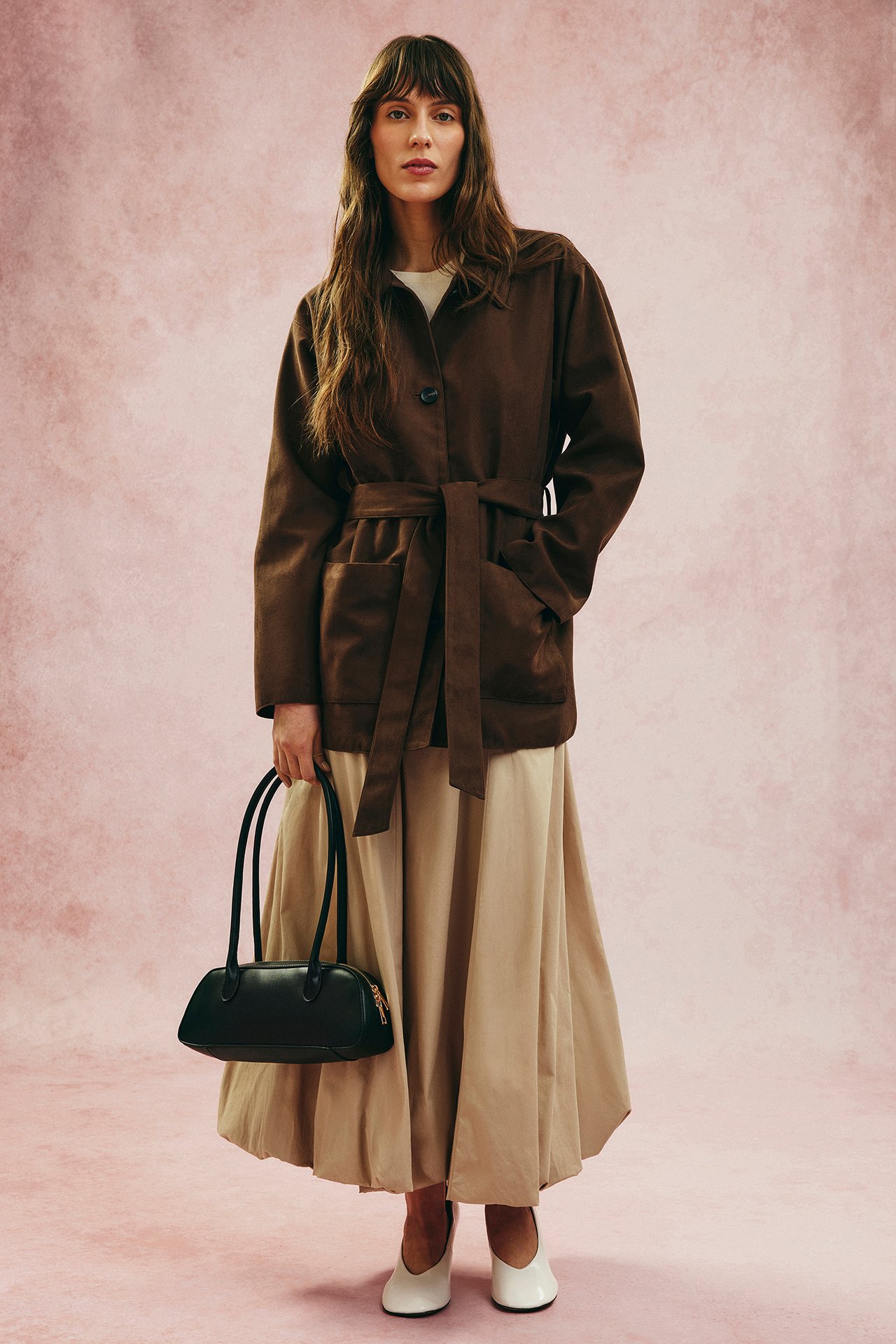 Frontutsikt: Kvinne i brun imitert semsket jakke med belte, beige skjørt, hvit topp, svart veske, hvite sko.