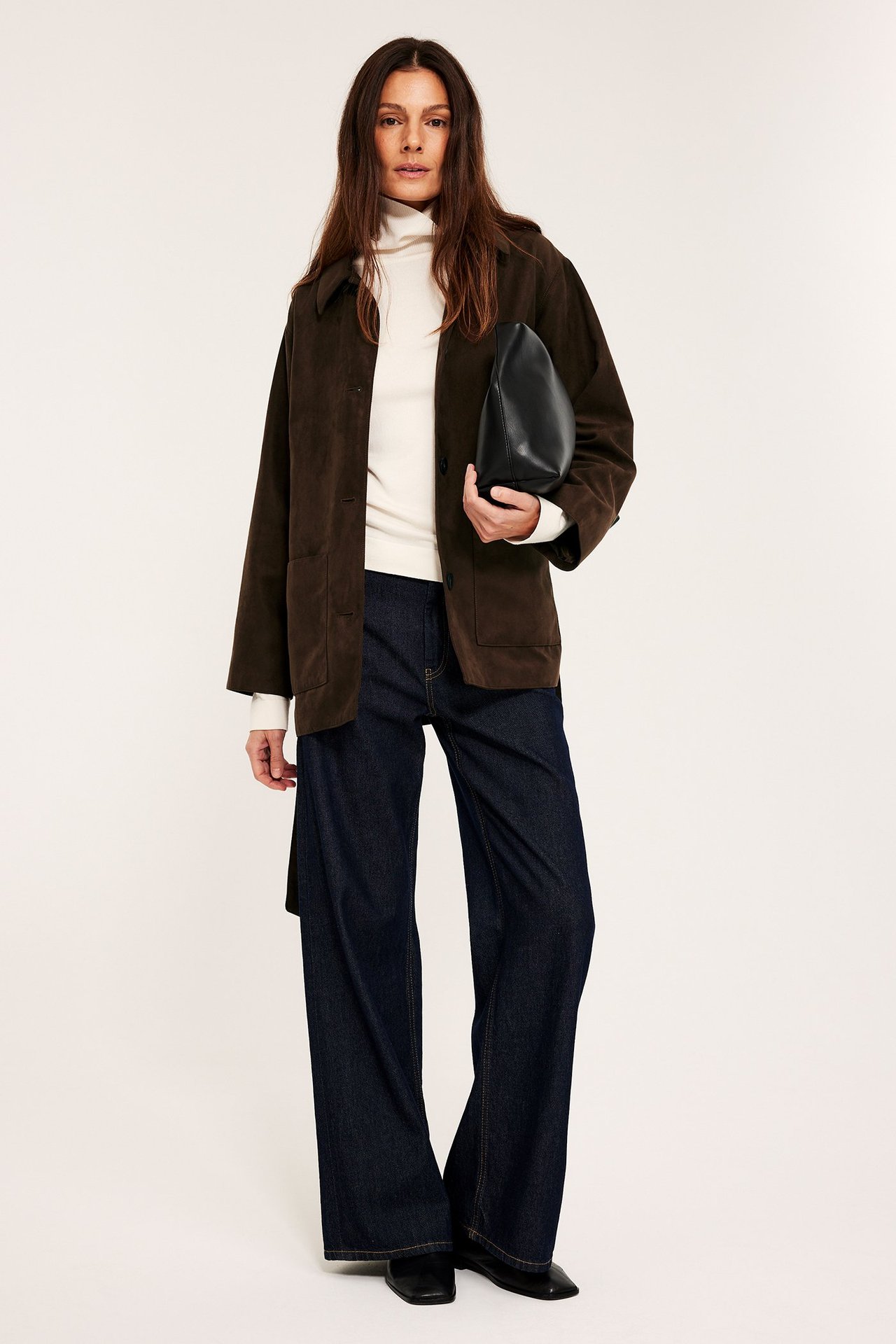 Etukuvassa: Nainen pukeutuneena avonaiseen ruskeaan mokkajäljitelmätakkiin, kermaan poolopaitaan, tummansinisiin farkkuihin, pitää mustaa clutchia.