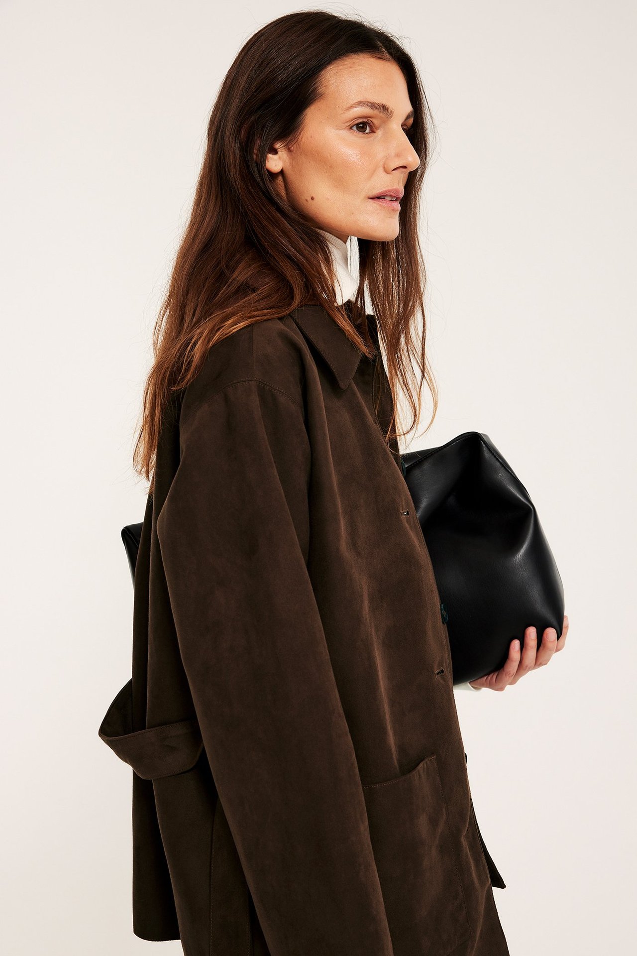 Sidovy av kvinna i brun jacka i mockaimitation med bälte, vit topp, håller svart handväska.