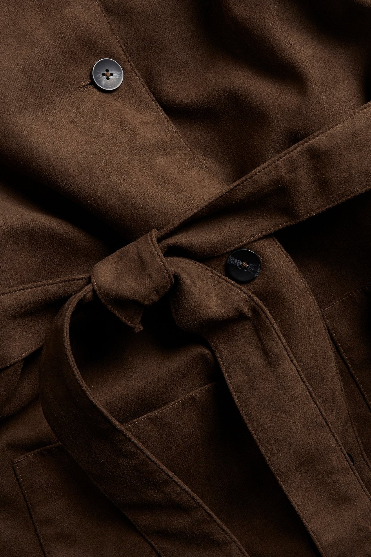 Närbild av en brun jacka i mockaimitation med knutet bälte och mörka knappar.