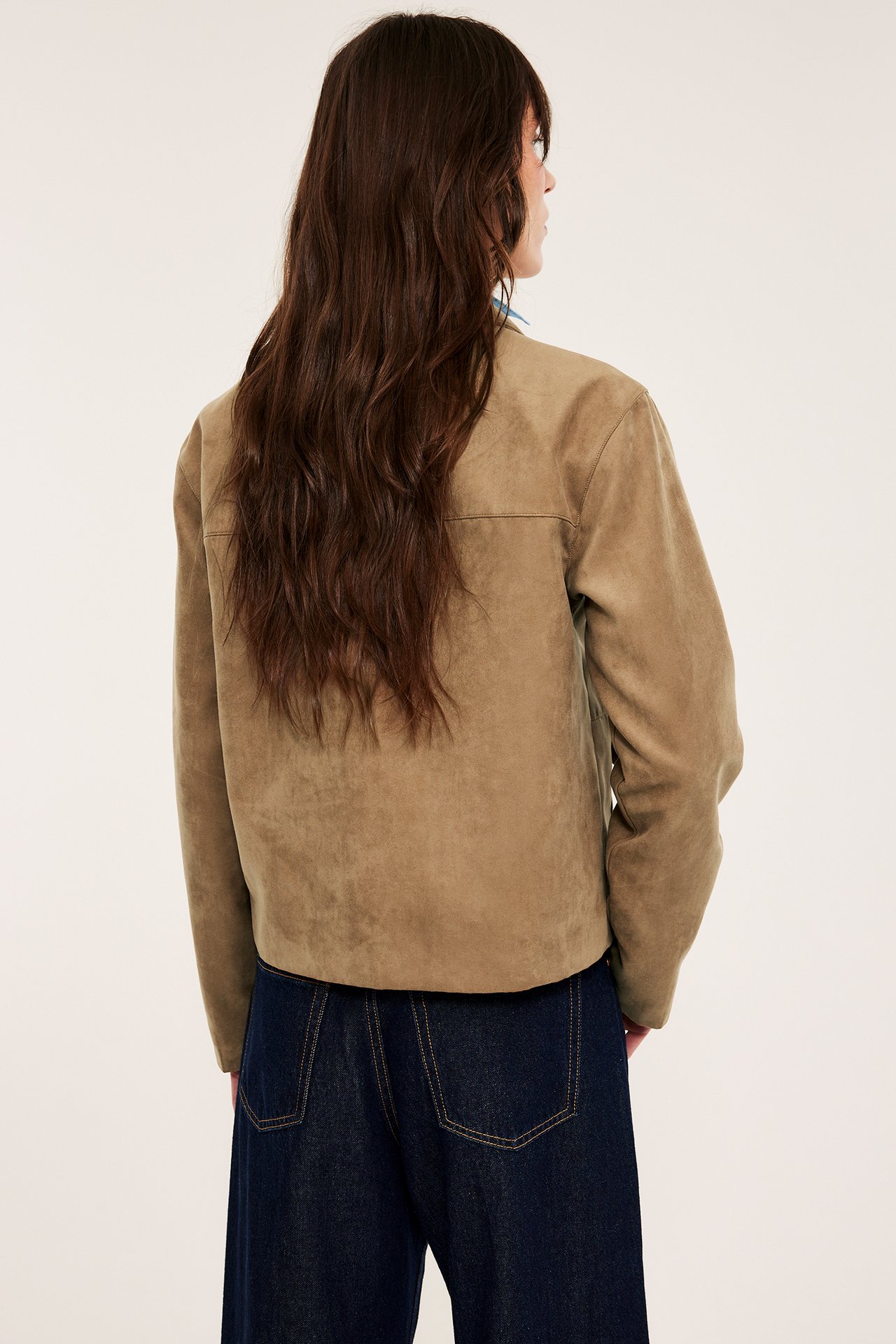 Kvinne bakfra i brun semsket jakke over blå topp med mørkeblå jeans.