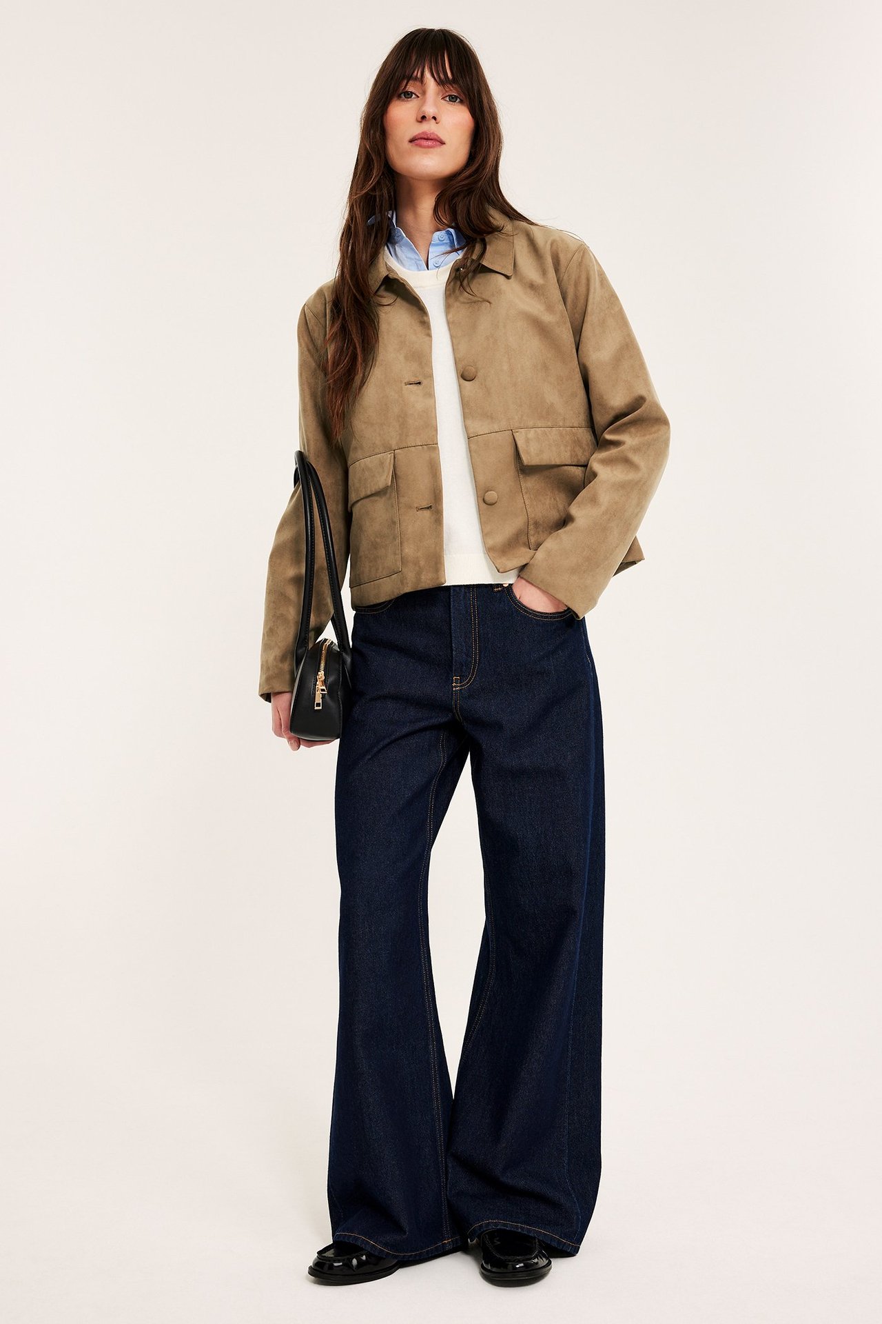 Widok z przodu: Kobieta w brązowej kurtce zamszowej, ciemnoniebieskich jeansach, czarnych mokasynach, z czarną torebką.