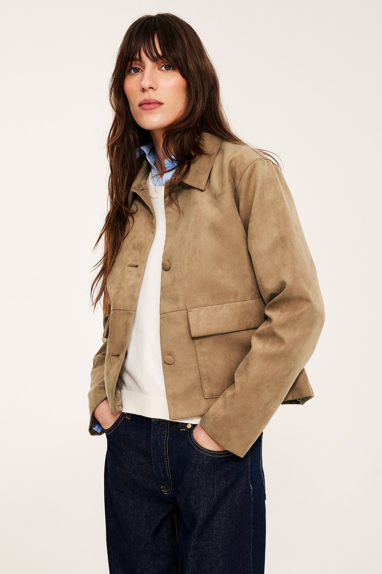 Kobieta w brązowej zamszowej kurtce z przednimi kieszeniami, na białym swetrze i koszuli.