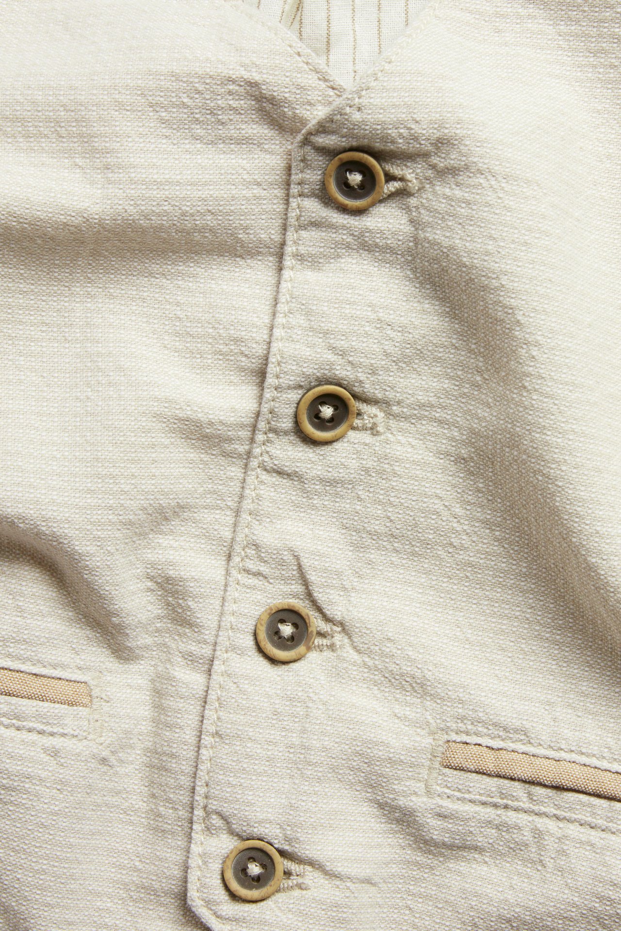 Närbild: Beige vävd bomullsväst med bruna knappar och dekorativa fickor; randigt foder synligt.