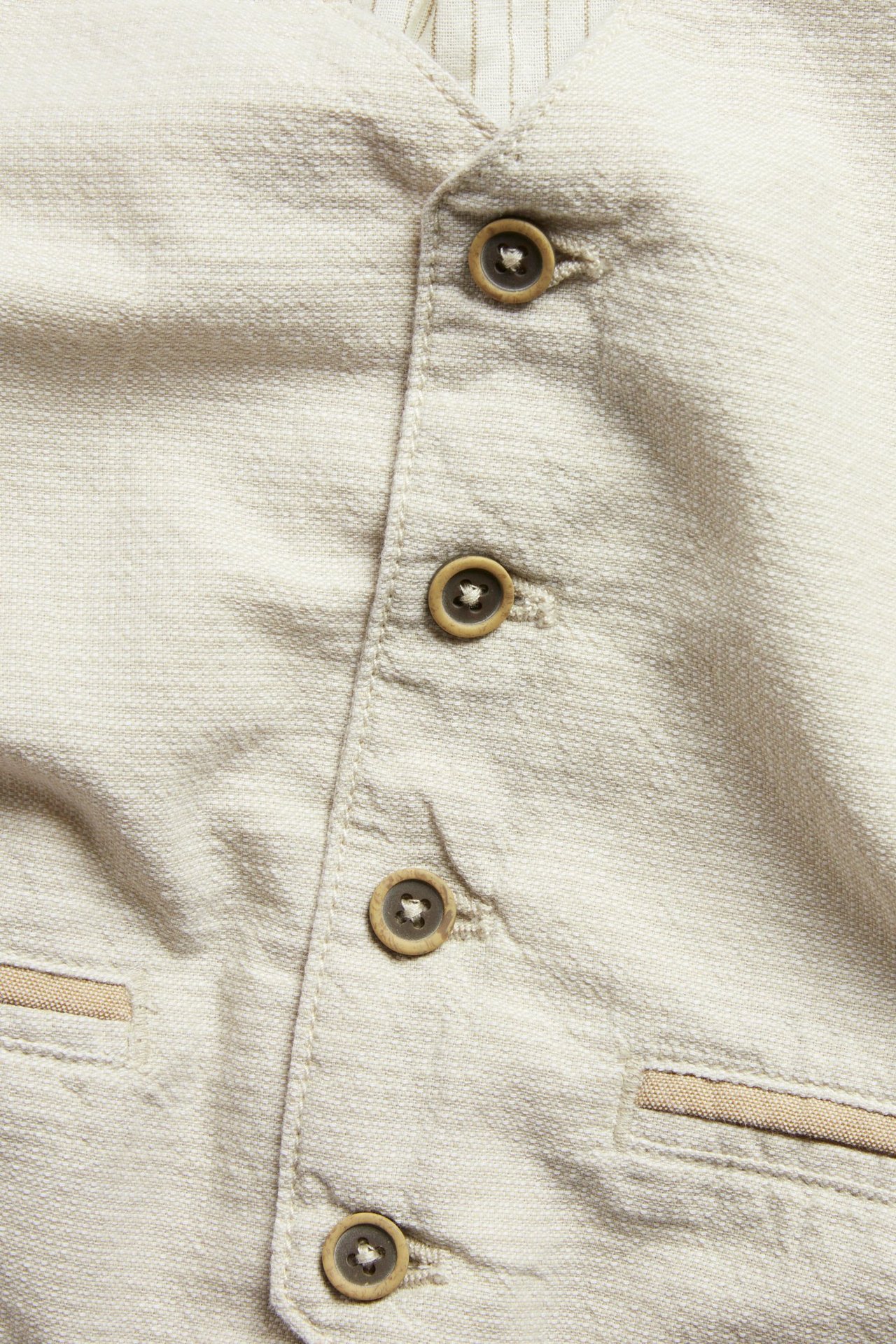Zbliżenie: Beżowa tkana bawełniana kamizelka z brązowymi guzikami i ozdobnymi kieszeniami; widoczna podszewka w paski.