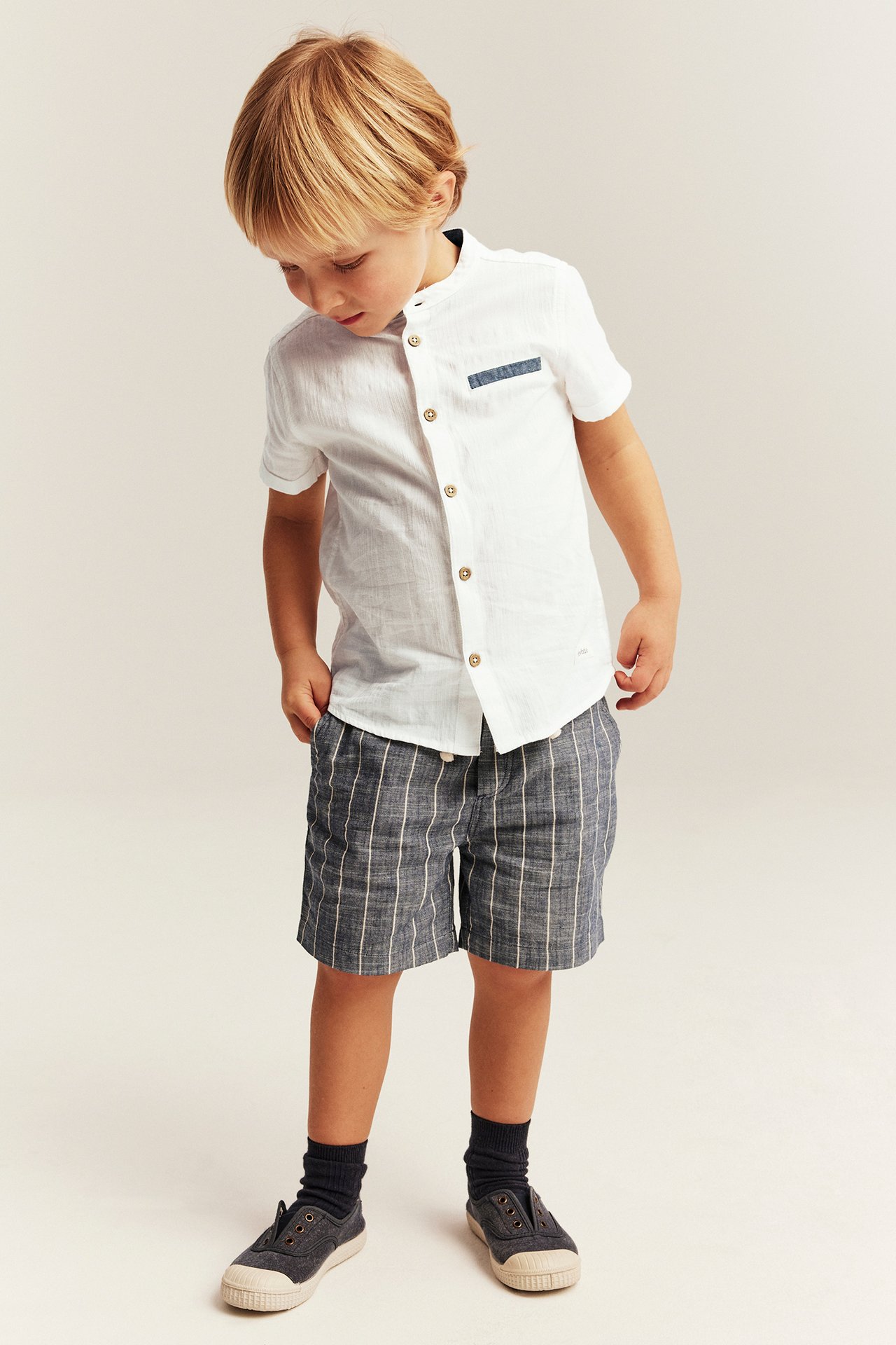 Forfra, et barn i en offwhite kortermet vevd skjorte, mørkeblå rutete shorts, mørke sokker og joggesko.