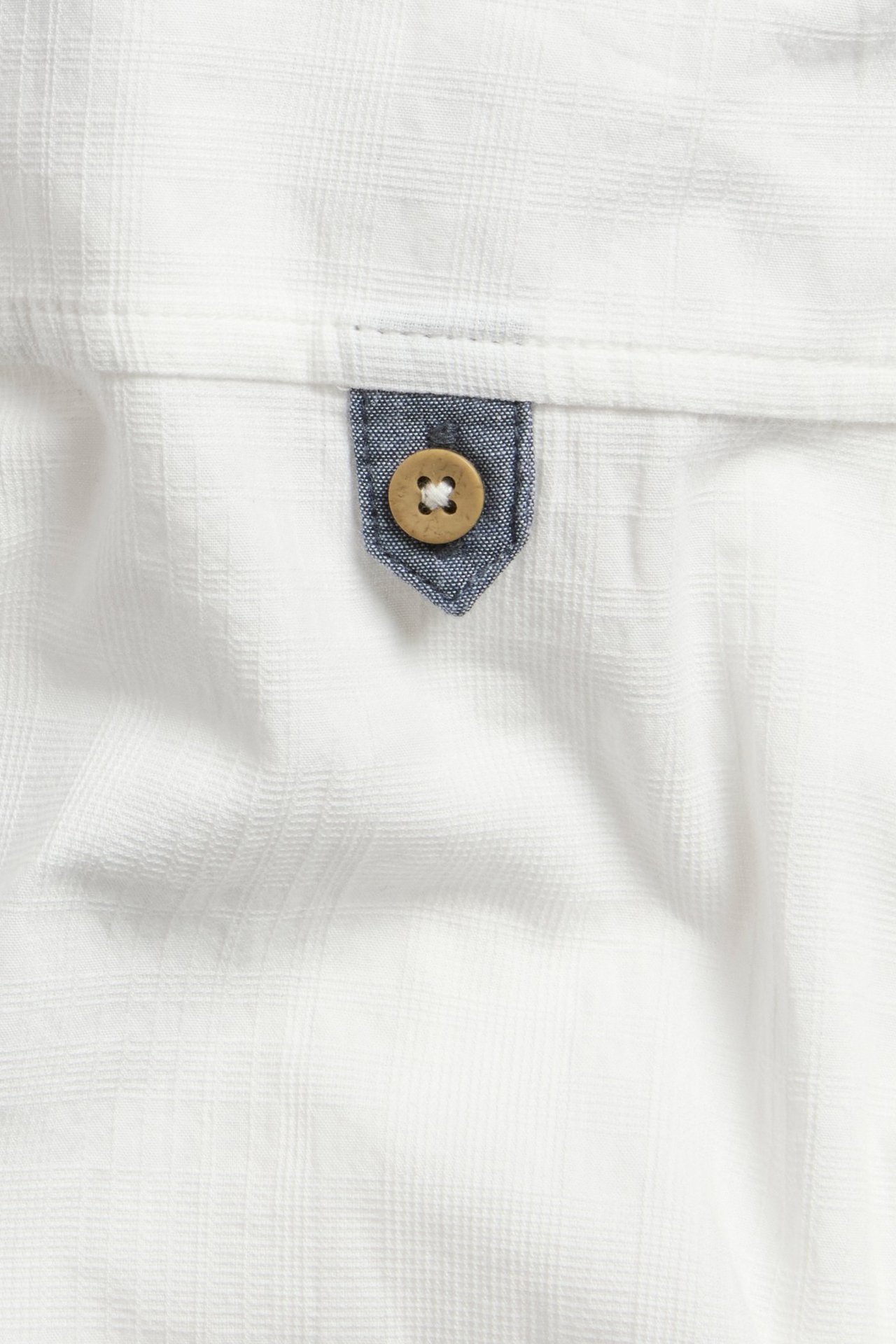 Zbliżenie na kremową bawełnianą koszulę, z subtelną kratką, niebieską patką chambray i drewnianym guzikiem.