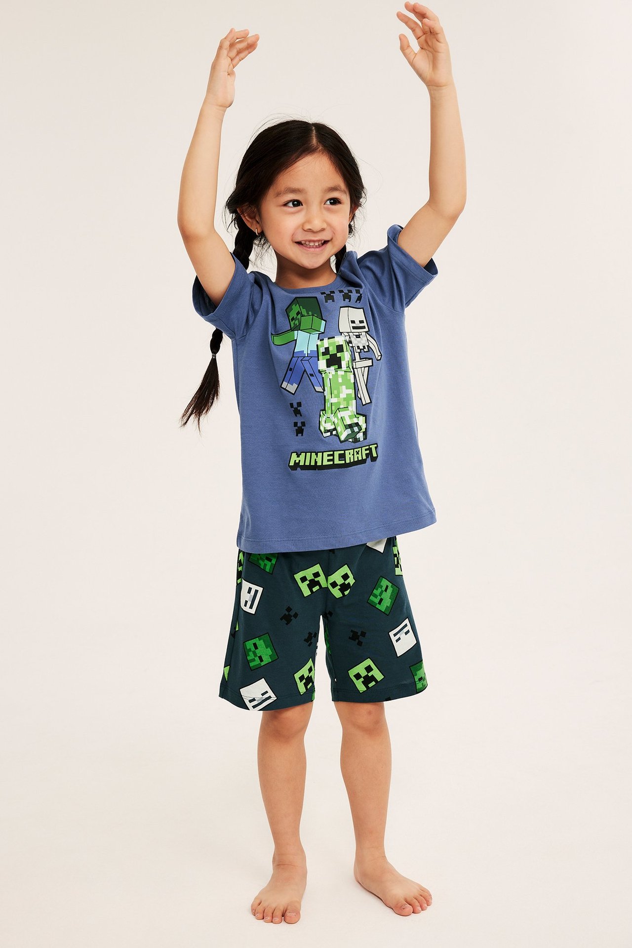 Widok z przodu: dziecko w niebieskiej piżamie Minecraft: koszulka z nadrukiem i ciemnozielone spodenki we wzory.