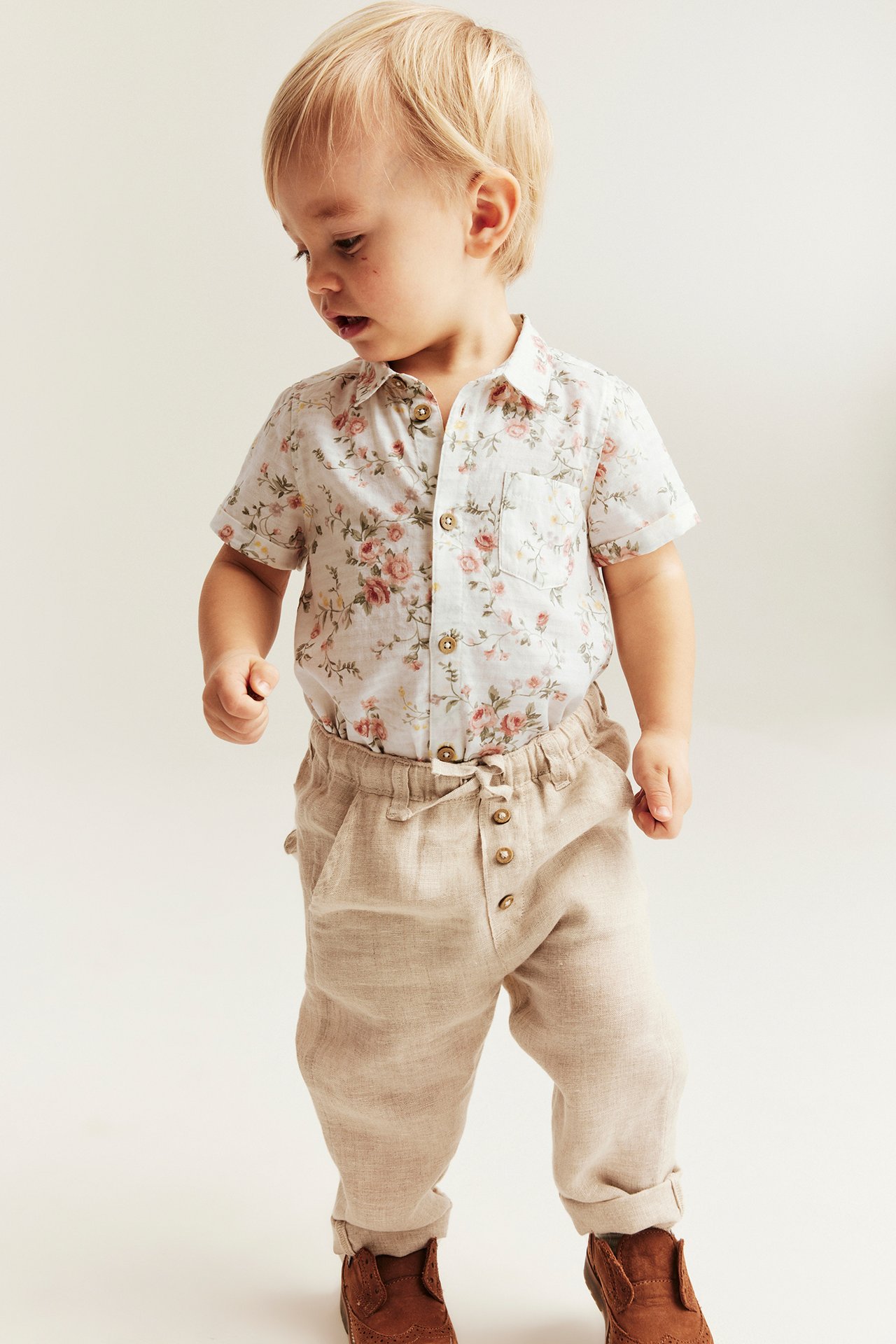 Frontalt bilde: baby i offwhite blomstret kortermet bomullsskjortebody og beige bukser.