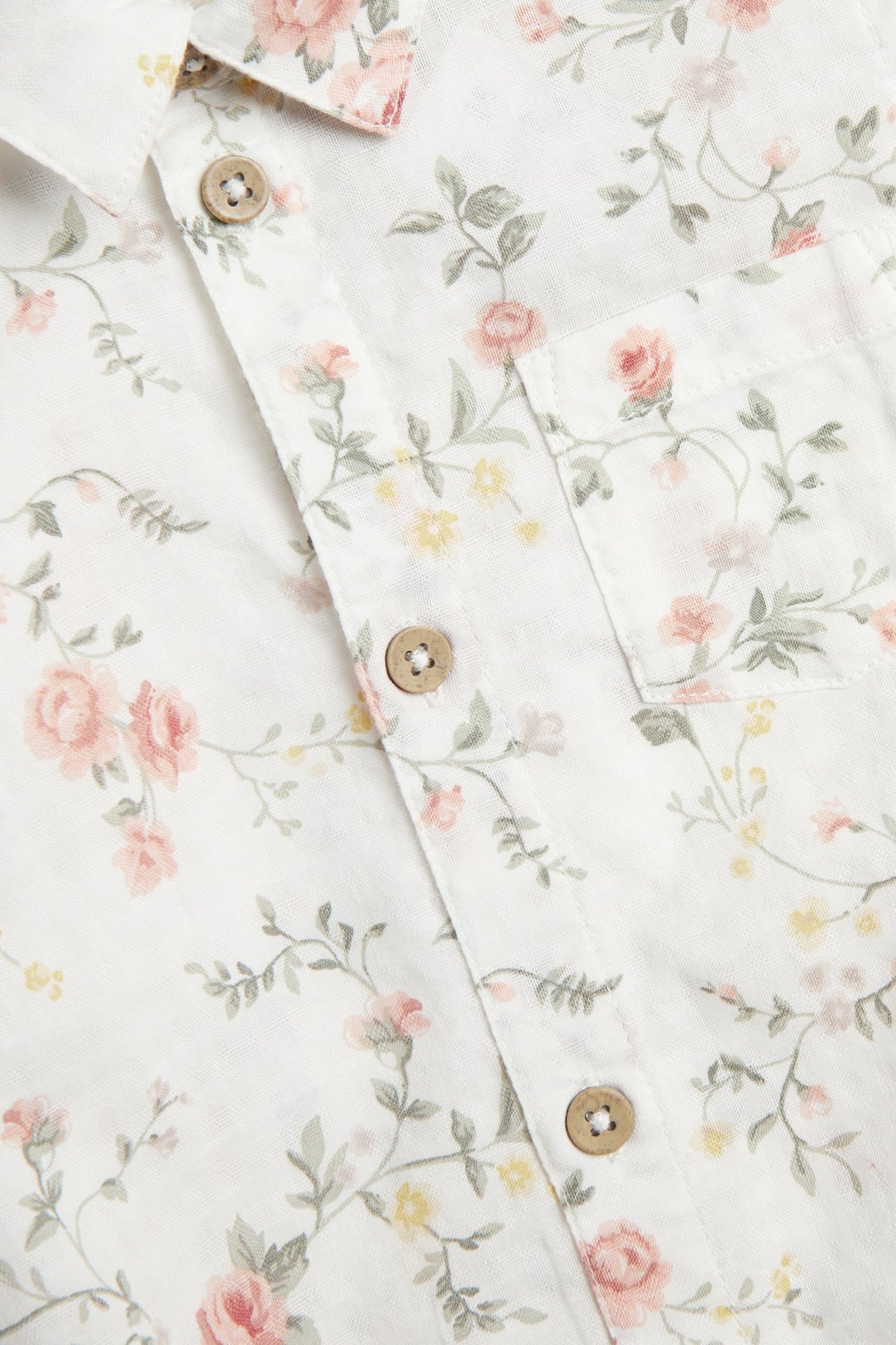 Närbild på en offwhite blommig kortärmad baby skjortbody med träknappar fram och en bröstficka.