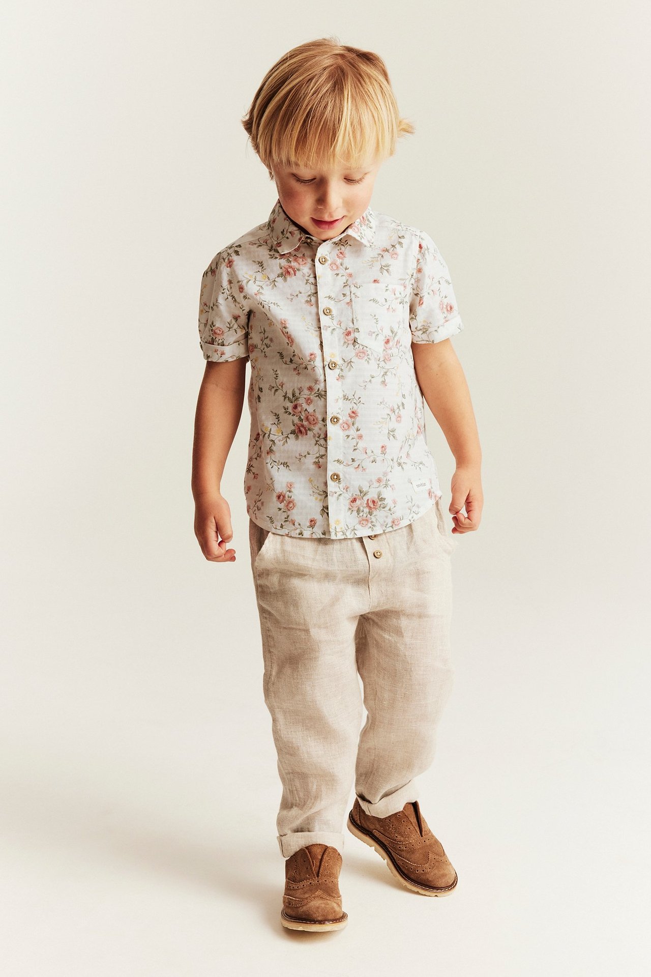 Widok z przodu: dziecko w białej koszuli w kwiaty, jasnobeżowych spodniach i brązowych butach.