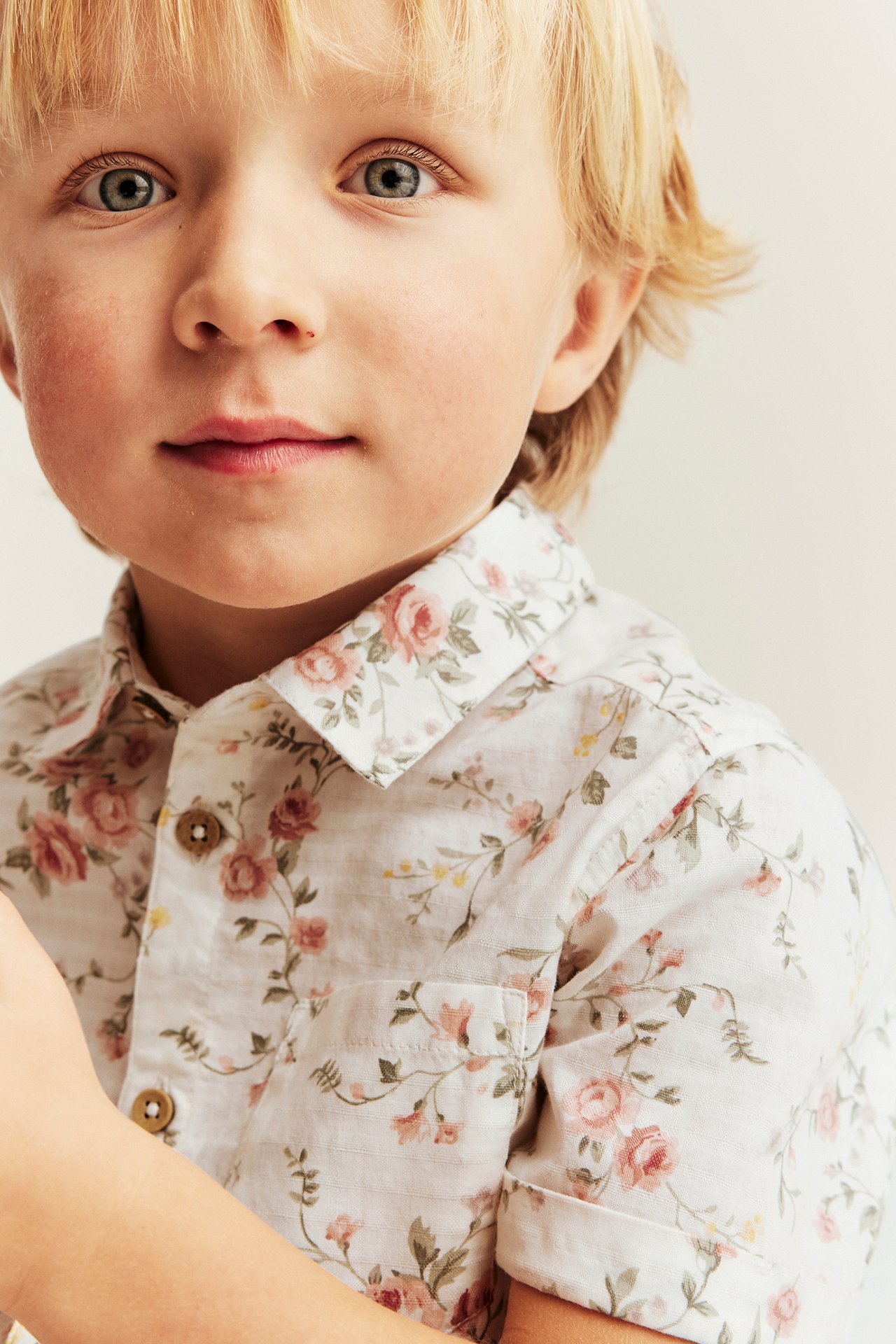 Lähikuva edestä lapsesta luonnonvalkoisessa lyhythihaisessa kukkapaidassa ruskeilla napeilla.