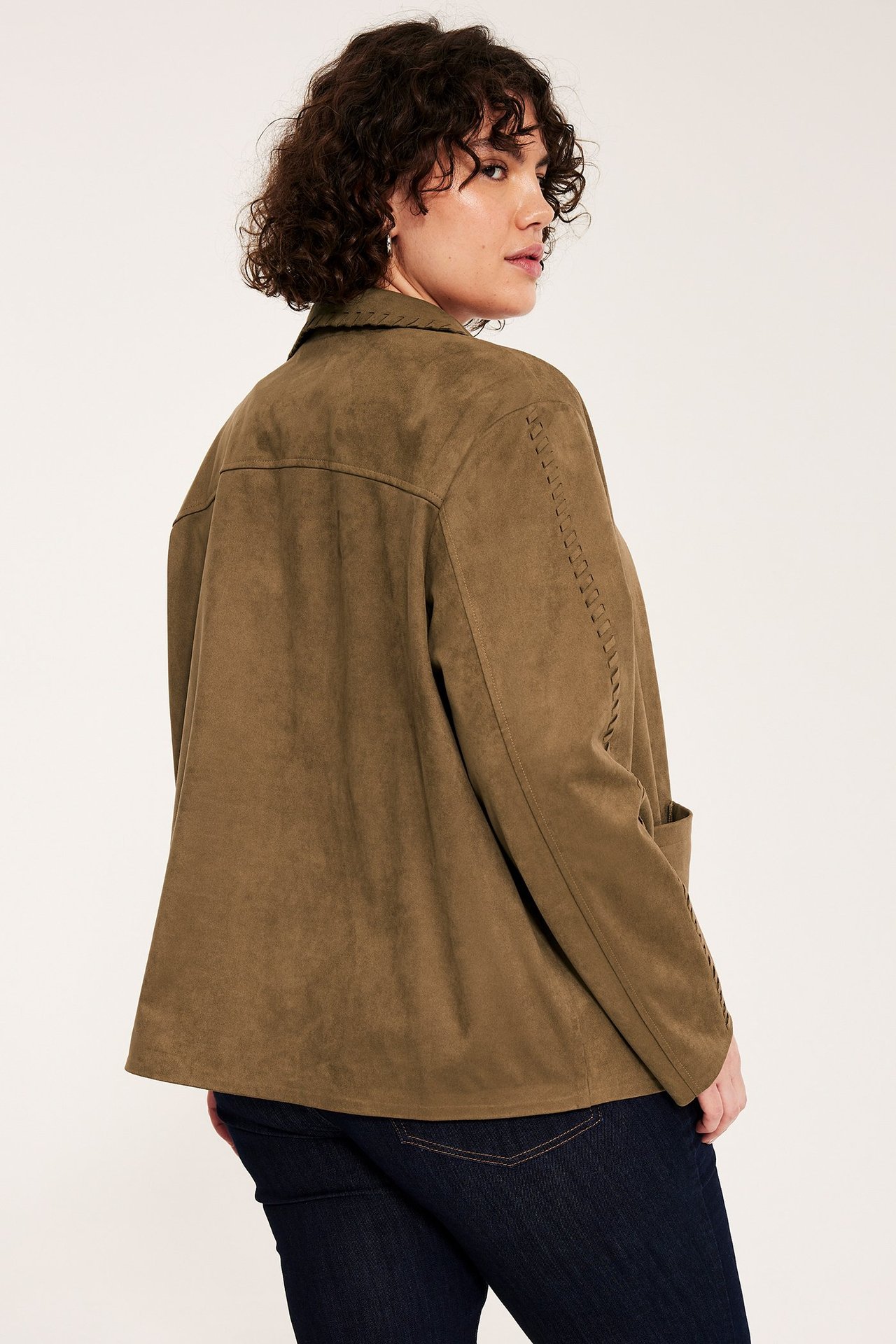 Rygg-/sida vy av en kvinna i en höftlång brun mockaimitationsjacka med dekorativa sömmar och mörkblå jeans.