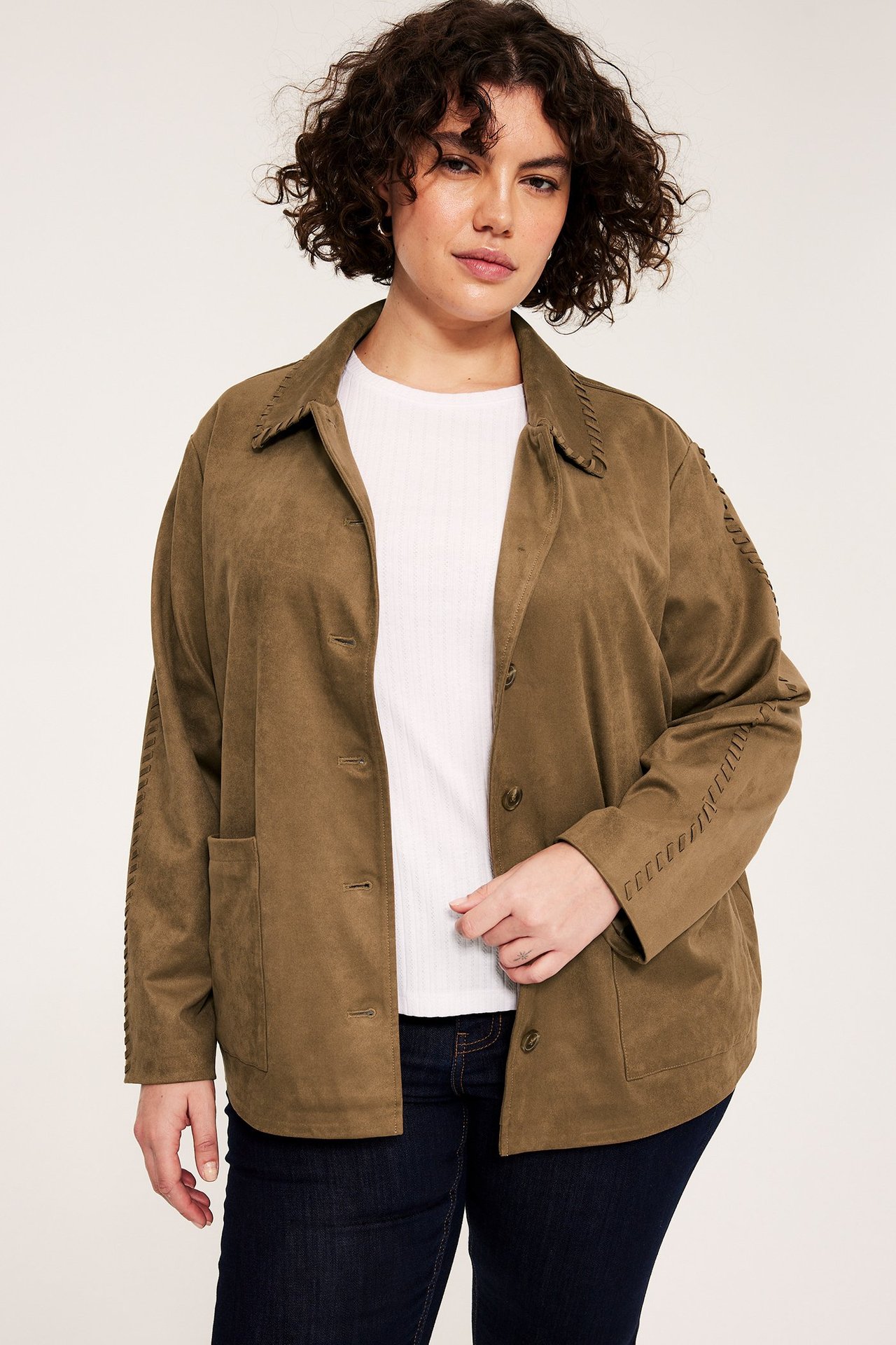 Front view av kvinna i khakibrun mockaimitationsjacka med dekorativa sömmar över vit topp och mörka jeans.