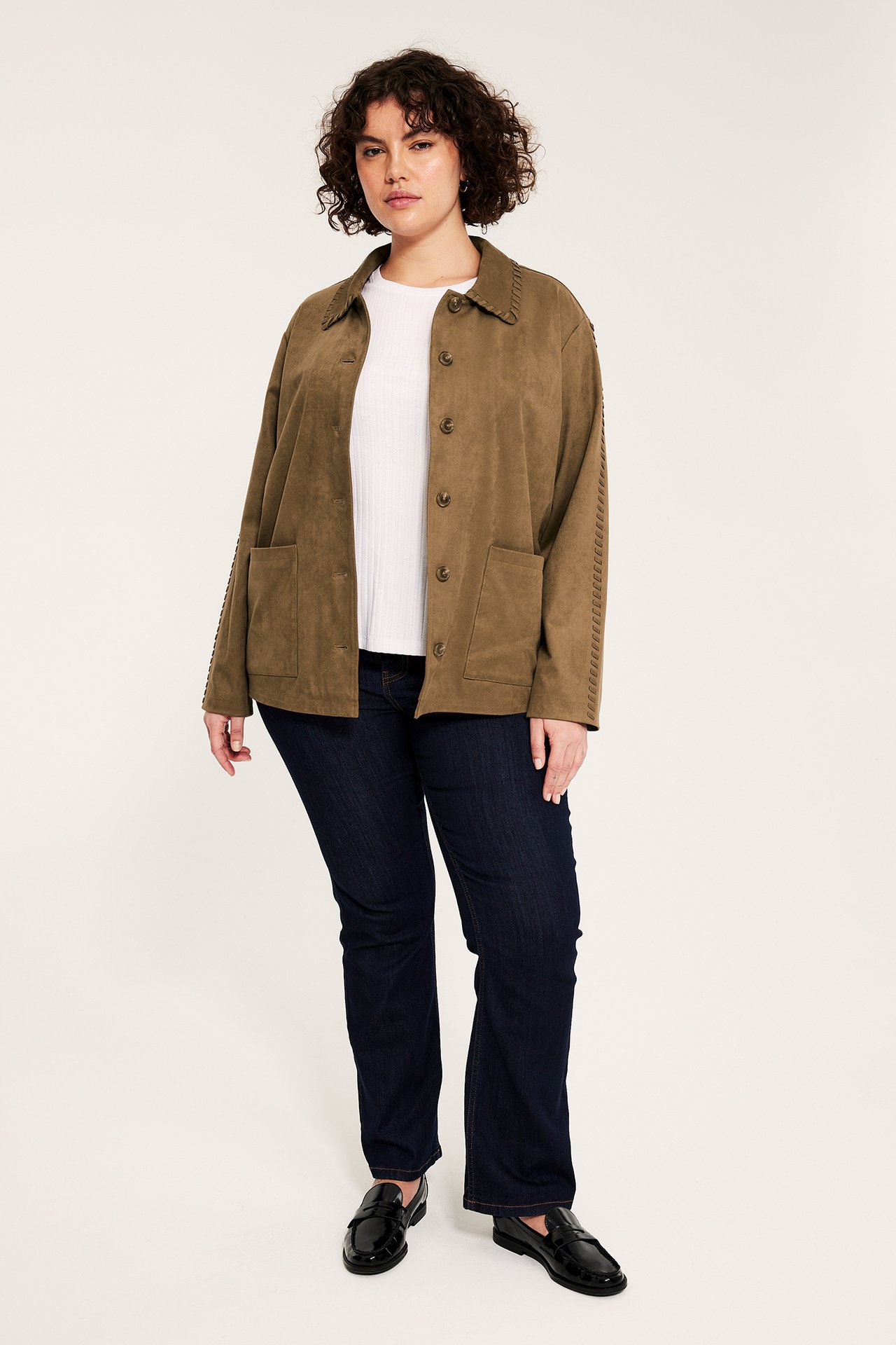 Framifrån: kvinna i brun jacka i mockaimitation med sömmar, vit t-shirt, mörka jeans, svarta loafers.