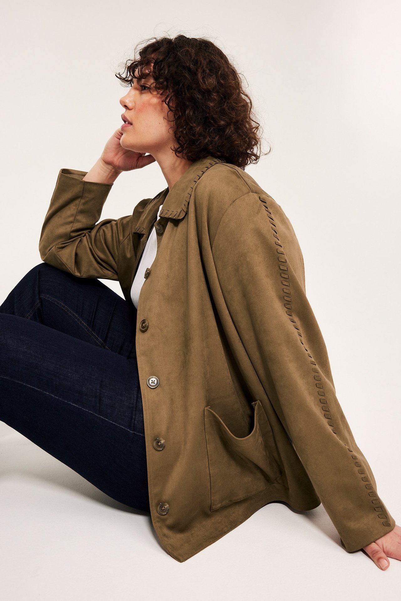 Widok z boku kobiety w brązowej kurtce z imitacji zamszu z ozdobnymi szwami i ciemnych dżinsach.