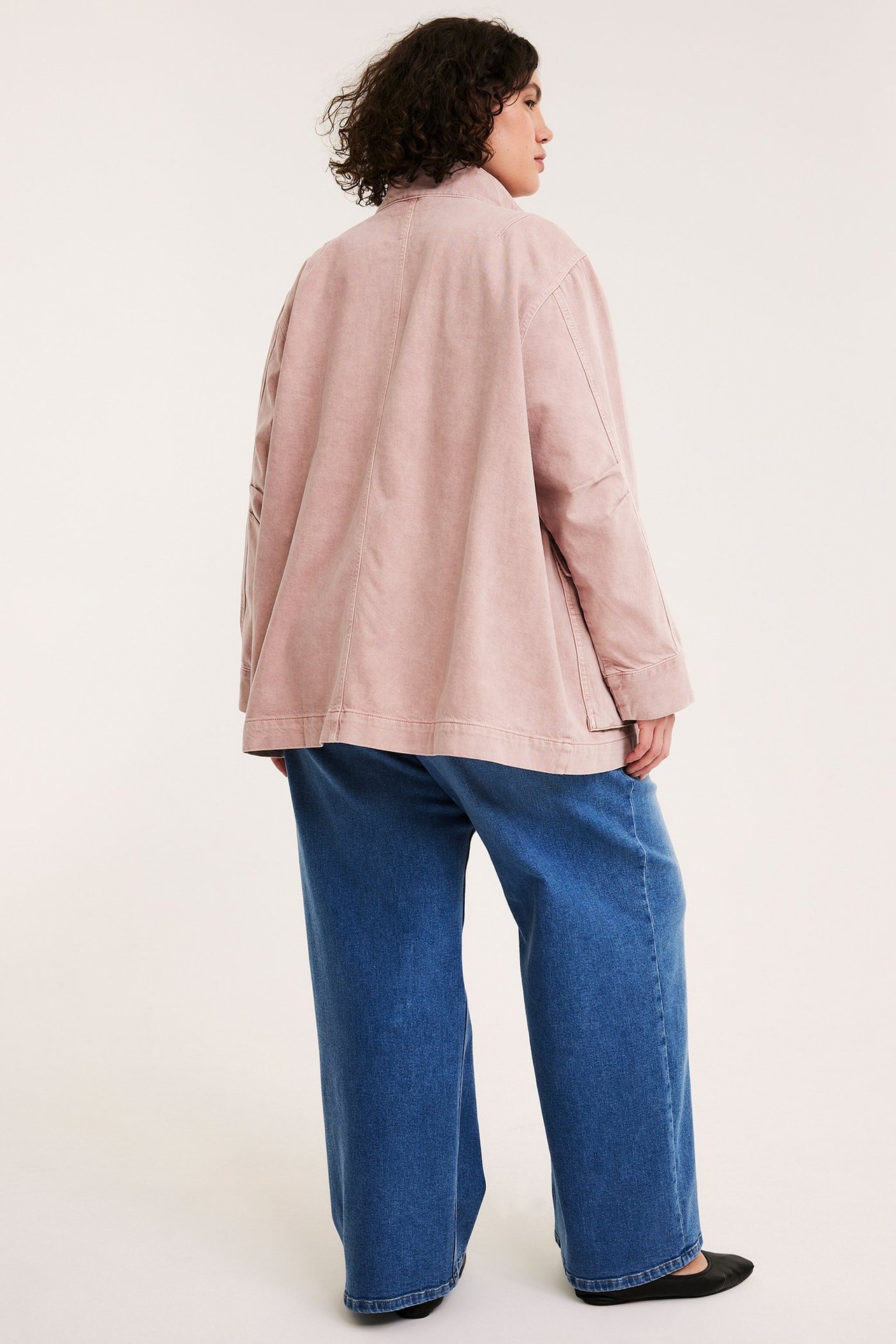 Naisen takasivukuva löysässä vaaleanpunaisessa puuvillatakissa, jossa on pystyskaulus, ja leveissä sinisissä farkuissa.