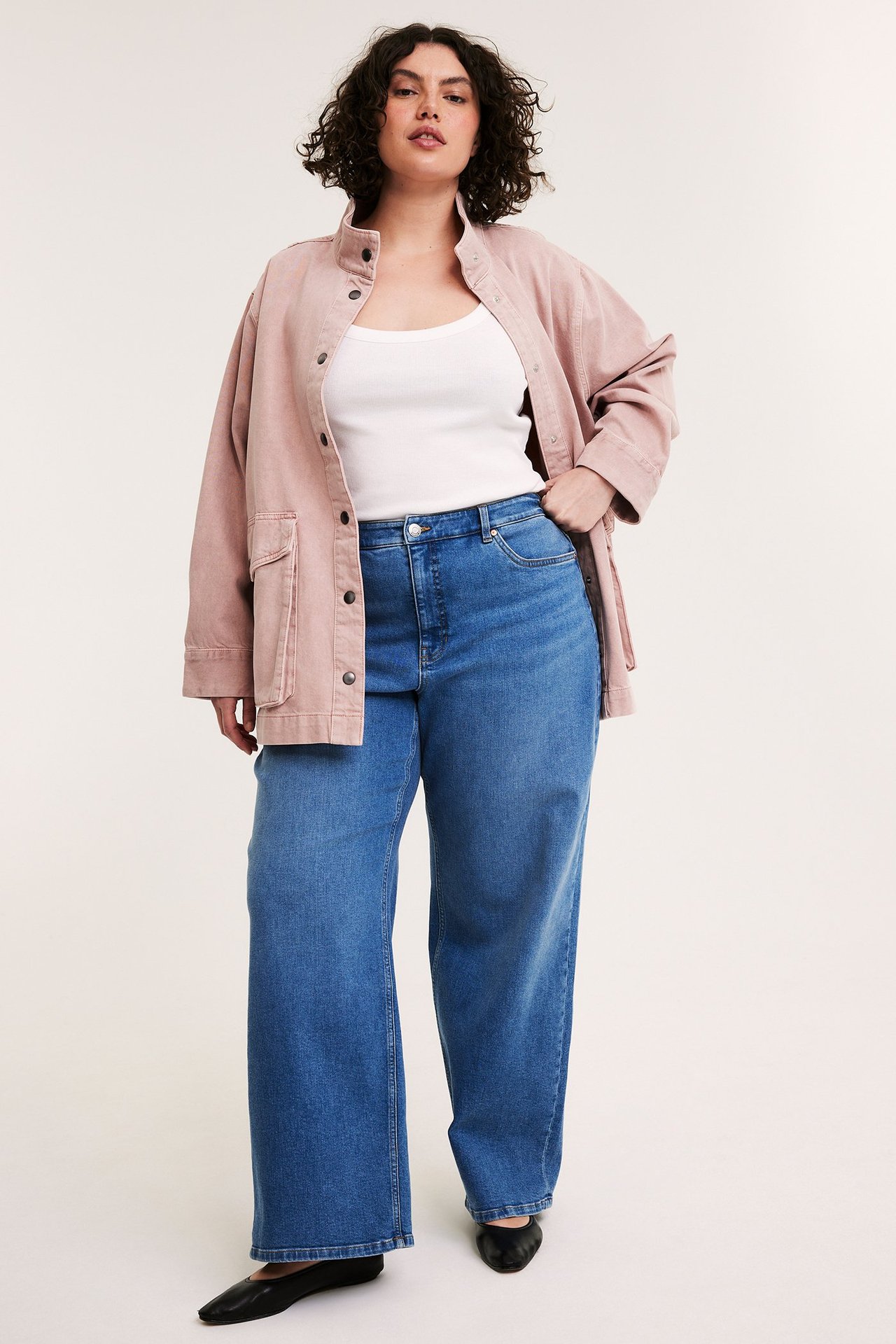 Kvinne i rosa bomullsjakke med store lommer, hvit singlet, blå jeans og svarte ballerinasko.