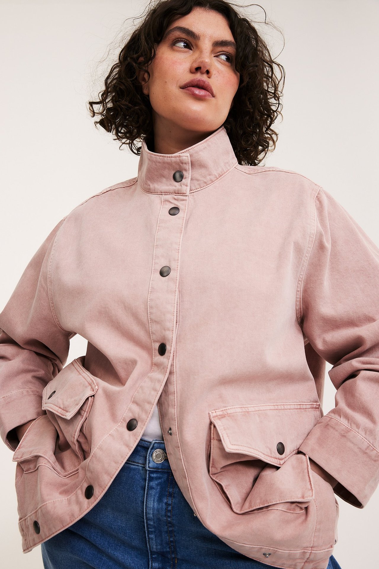 Nainen vaaleanpunaisessa puuvillatakissa, korkea kaulus, suuret taskut, tummat napit, sinisten farkkujen päällä. Edestä.