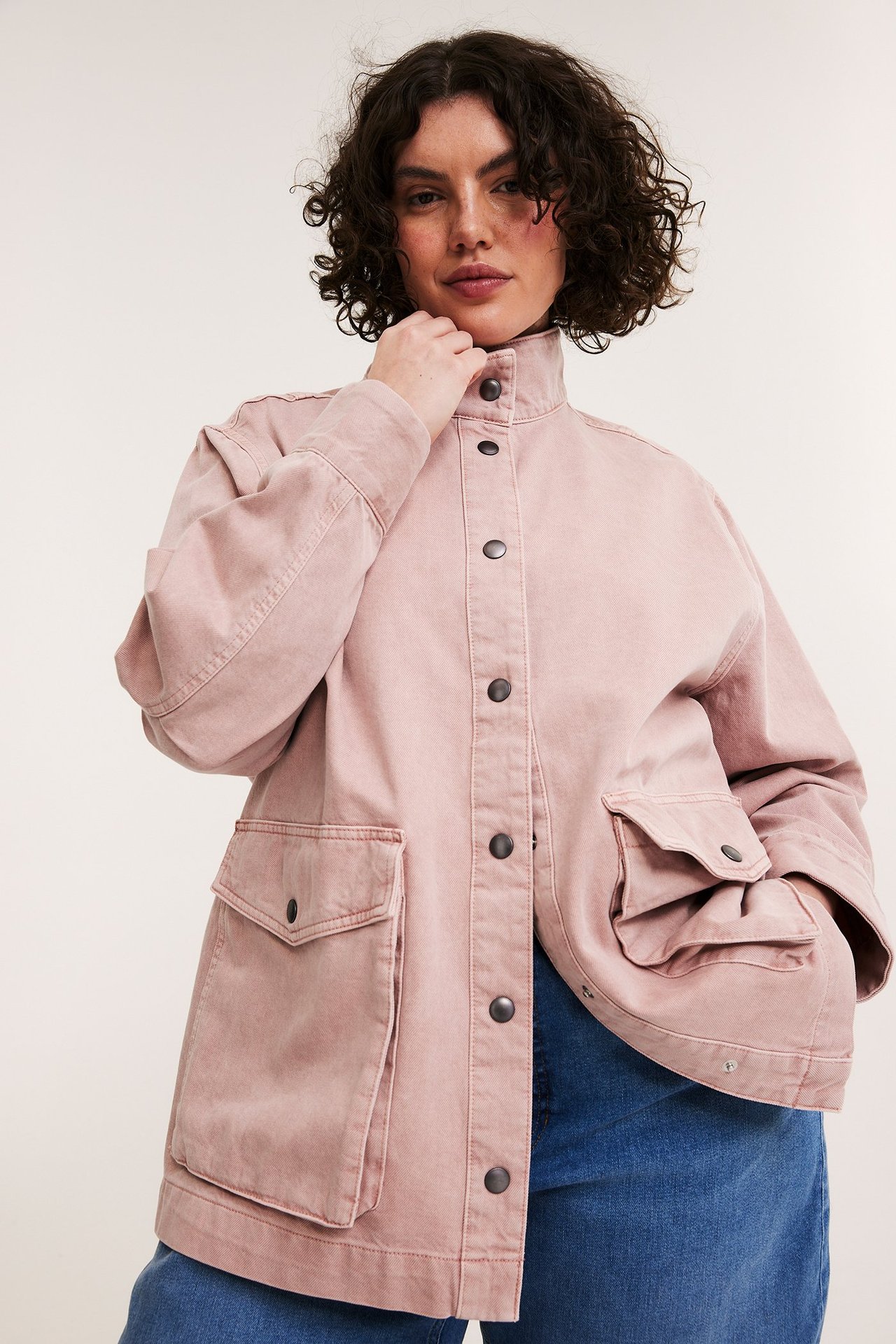 Widok z przodu: kobieta w różowej bawełnianej kurtce z dużymi kieszeniami, stójką, na niebieskich dżinsach.