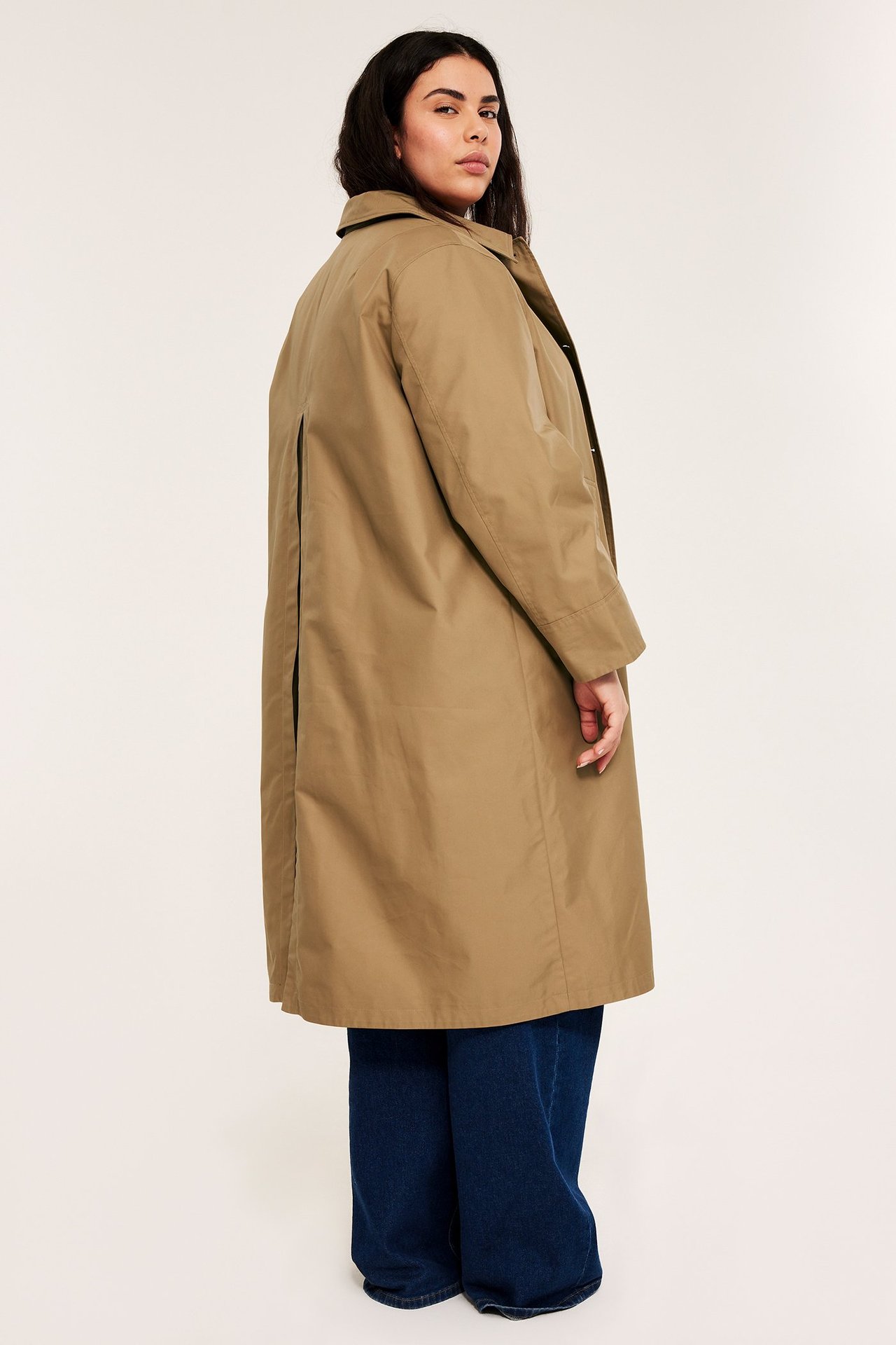 Nainen takaa ja sivulta: beigessä carcoatissa selkävekeillä, tummansinisten leveälahkeisten farkkujen päällä.