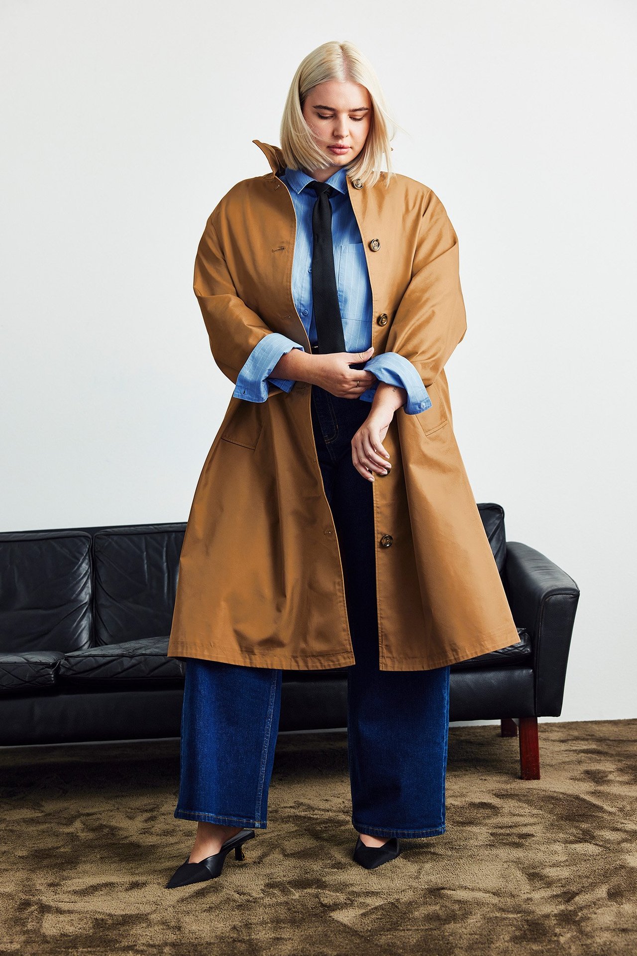 Etukuva: Nainen löysässä beigessä takissa, sinisessä paidassa, mustassa solmiossa, tummansinisissä farkuissa, mustissa koroissa.