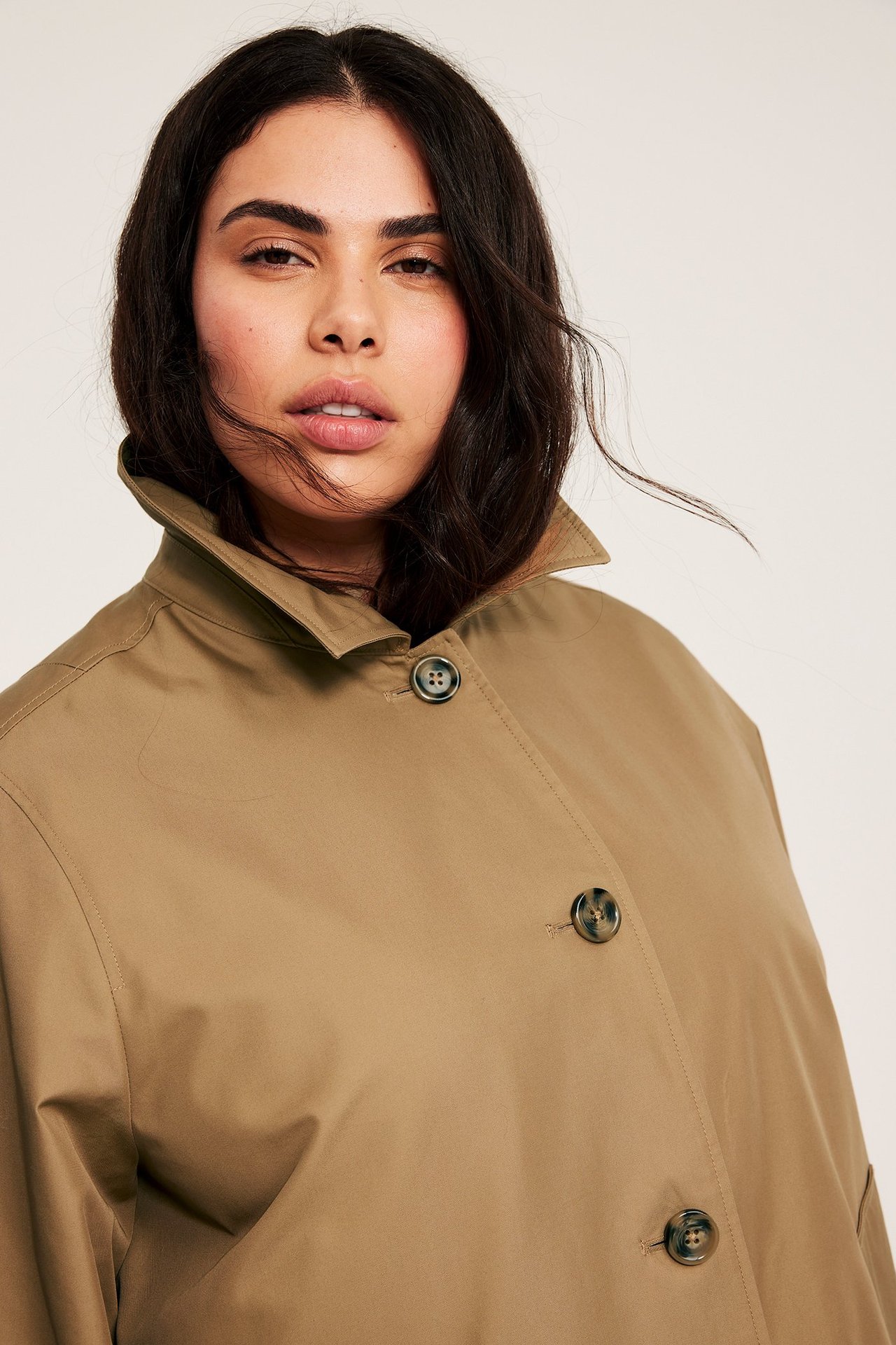 Lähikuva edestä naisesta beigessä yksivärisessä takissa, jossa kaulus ja tummat napit.