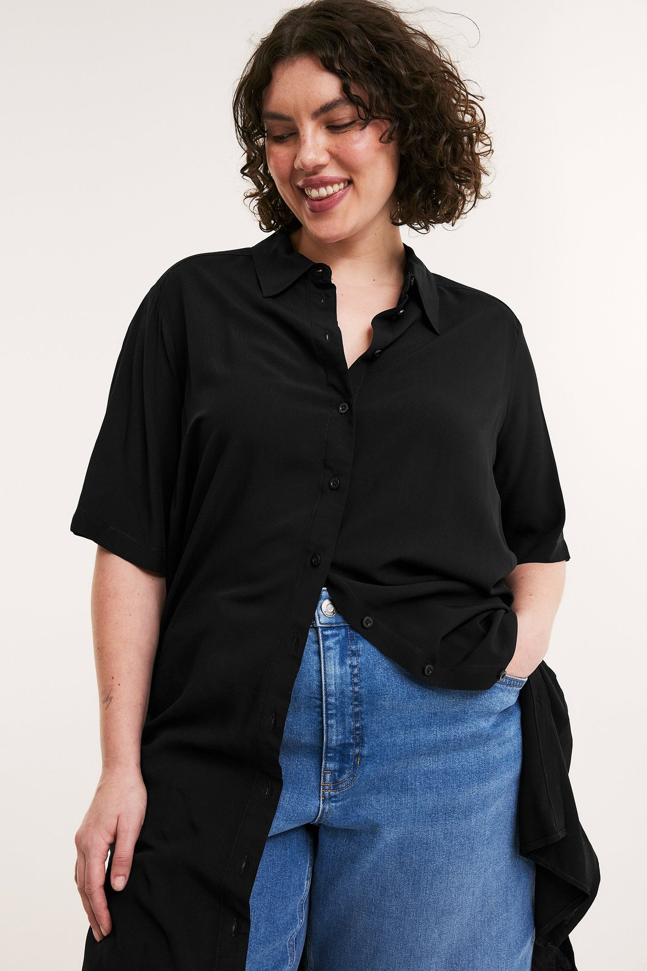 Edestä näkymä naisesta mustassa viskoosipaitamekossa avonaisena sinisten farkkujen päällä.