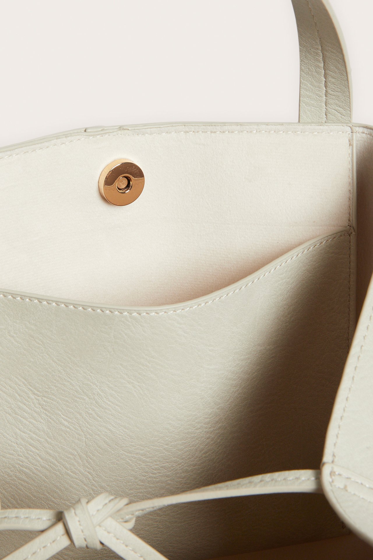 Lähikuva: vaalea beige tekonahkainen ostoskassi magneettisuljinnalla, sisätaskulla ja sidotulla hihnalla.