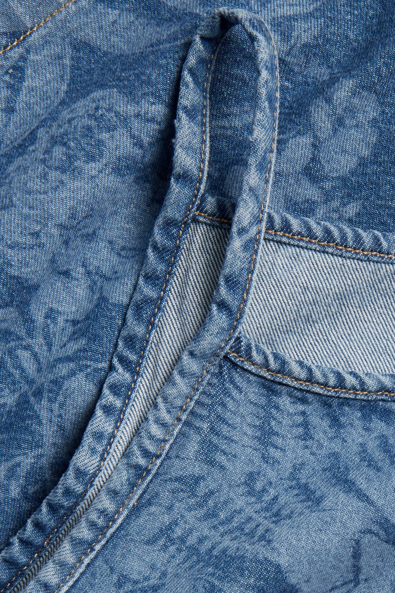 Närbild av ett ljust denimlinne som visar ett diskret blommigt mönster, en axelrem och sömdetaljer.