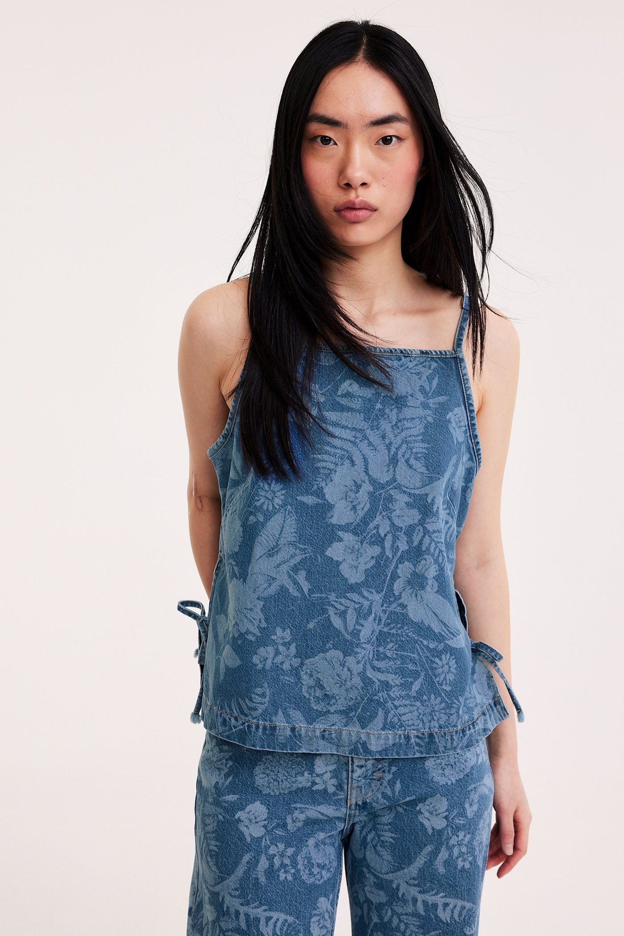 Naisen etukuva, yllään vaalea denimtoppi kukkakuviolla, halkioilla, solmionauhalla ja yhteensopivilla housuilla.