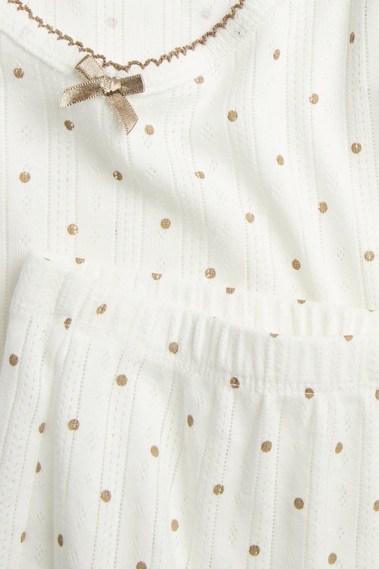 Nærbilde av et beige pyjamasett i bomullspointelle med gullprikker, bølgekant og sløyfe.
