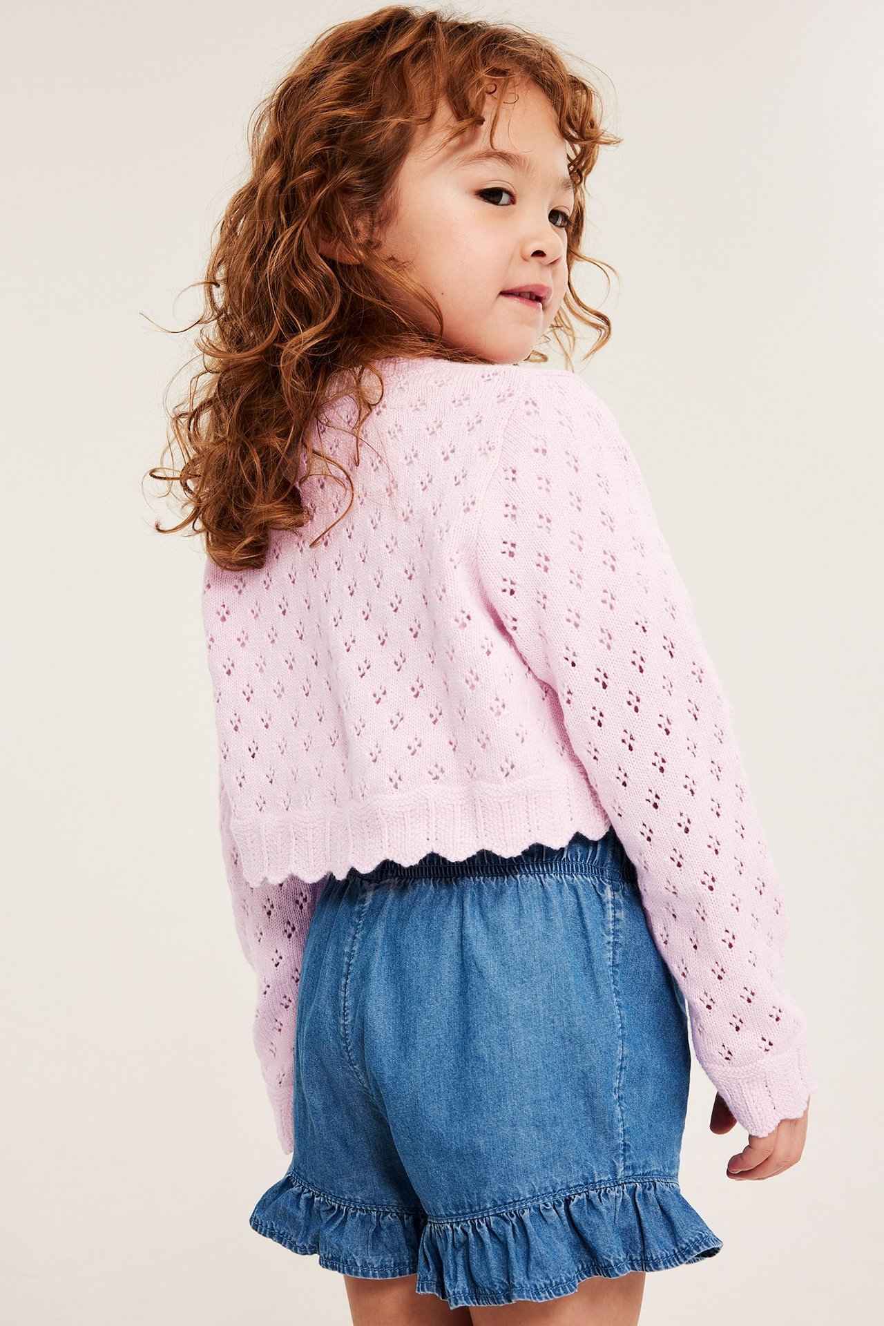 Bakvy av barn i ljusrosa mönsterstickad bomullskofta med vågig kant, över blå volangshorts.